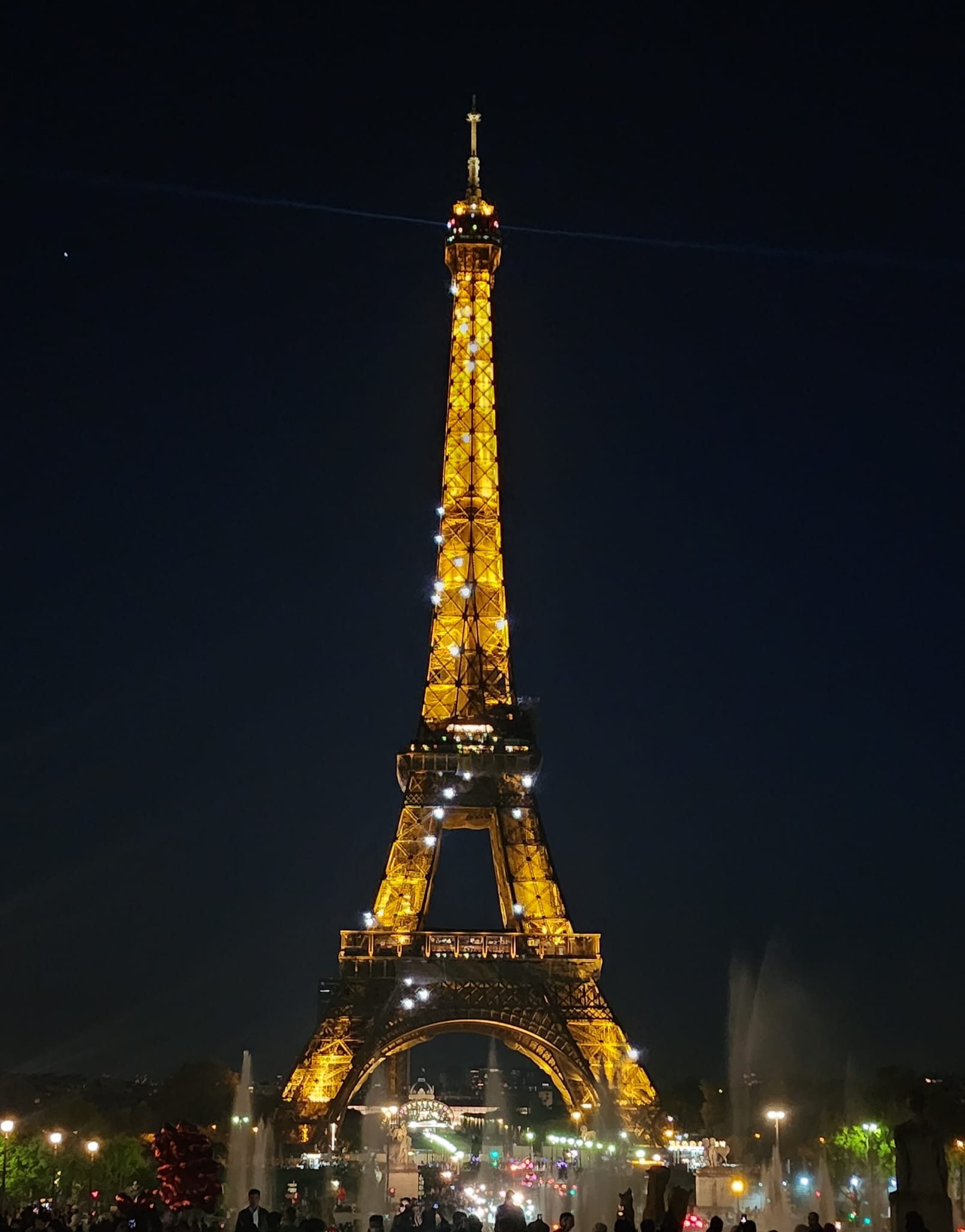 The Eiffel Tower brightly illuminated against the night sky, with sparkling lights dotting its structure, surrounded by a bustling crowd and fountains in the foreground.