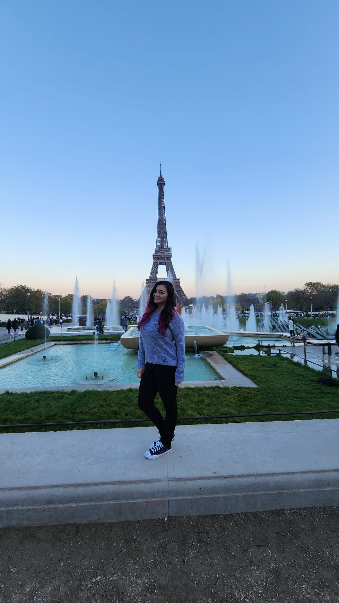 A person stands smiling in front of the Eiffel Tower, with fountains and green grass in the foreground, under a clear blue sky.
