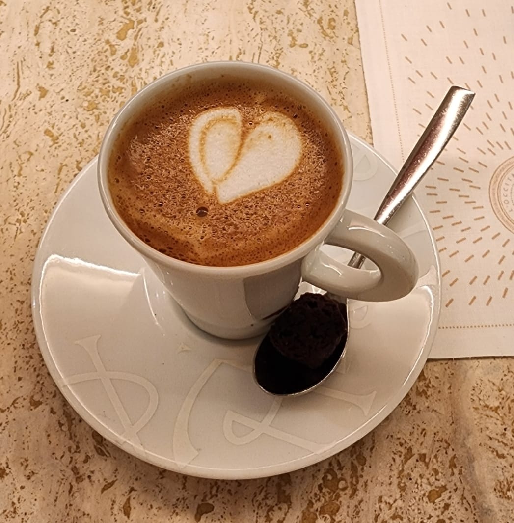 A cup of coffee with heart-shaped latte art sits on a white saucer, accompanied by a spoon holding a small chocolate treat, all placed on a marble table.