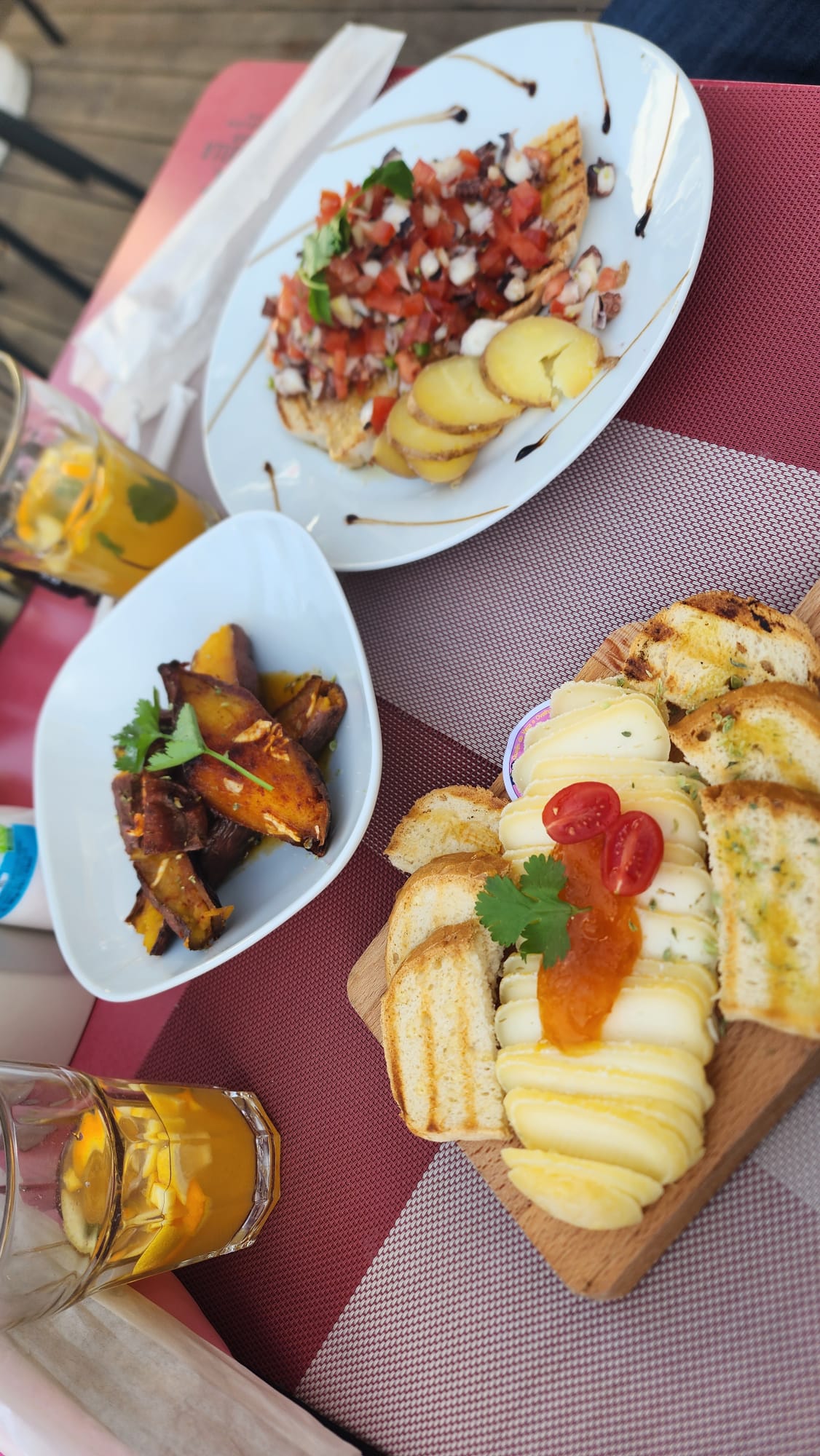 A table set with a variety of dishes, including a wooden board topped with sliced cheese, cherry tomatoes, and toasted bread. Nearby, a plate features grilled fish with diced tomatoes and herbs, accompanied by sliced potatoes. A bowl contains roasted sweet potato wedges, and glasses of a refreshing citrus drink are visible in the background.