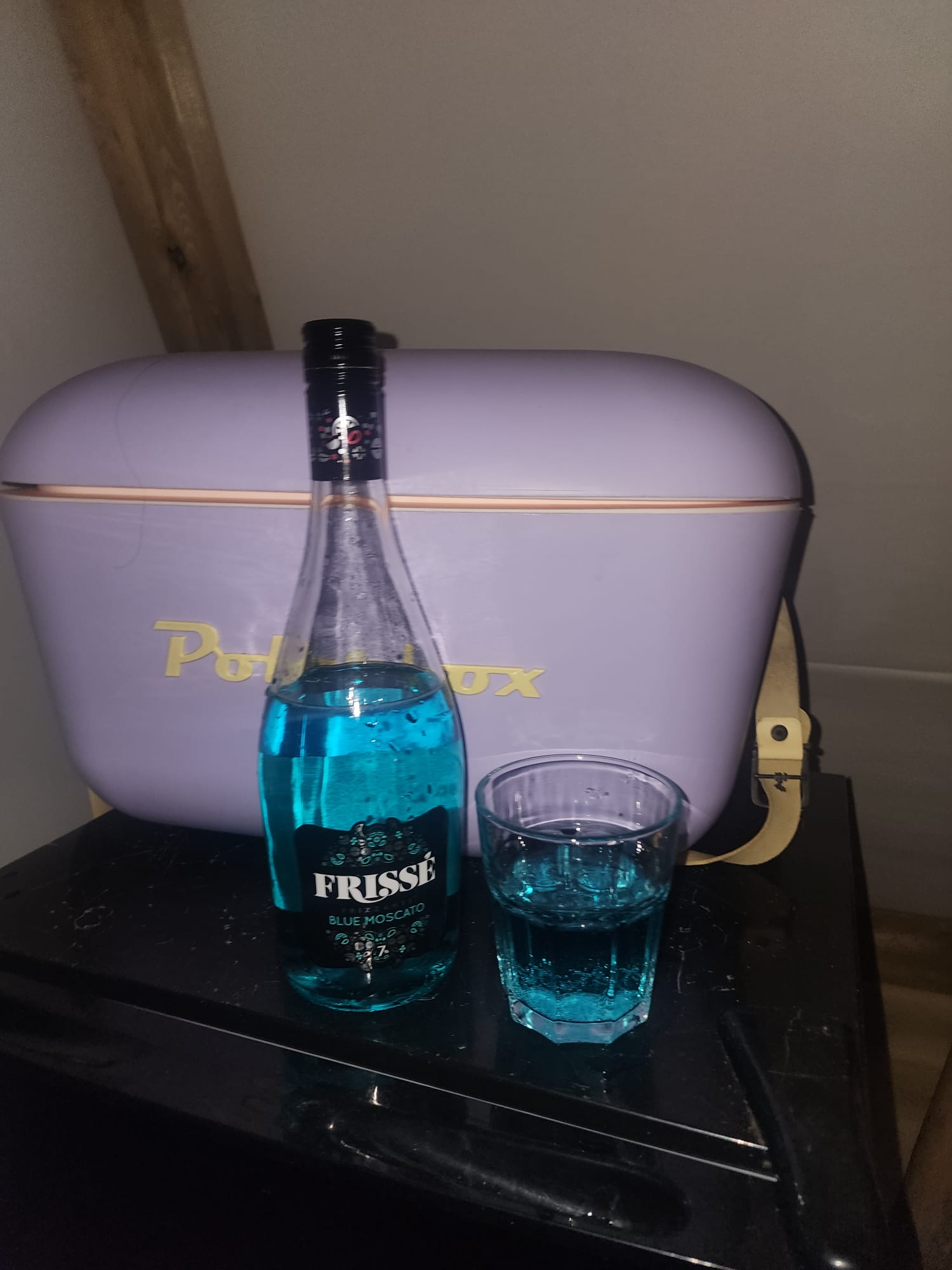 A bottle of Frissé Blue Moscato and a glass filled with the vibrant blue drink are placed on a black surface. Behind them is a pastel purple Polibox cooler with gold lettering.