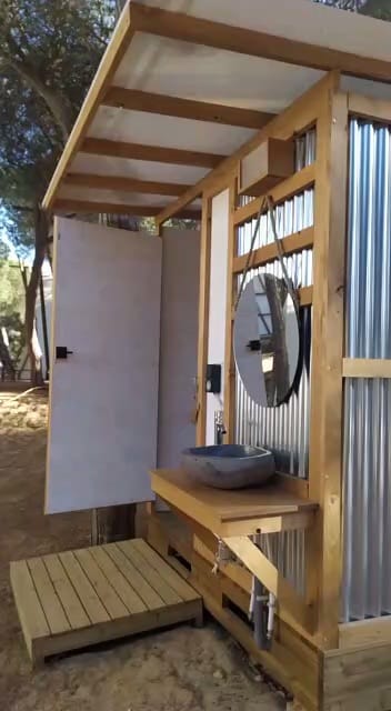 A rustic outdoor bathroom features a wooden structure with corrugated metal walls, a round mirror, and a stone basin sink. The open door reveals a simple interior, and the setting is surrounded by trees.