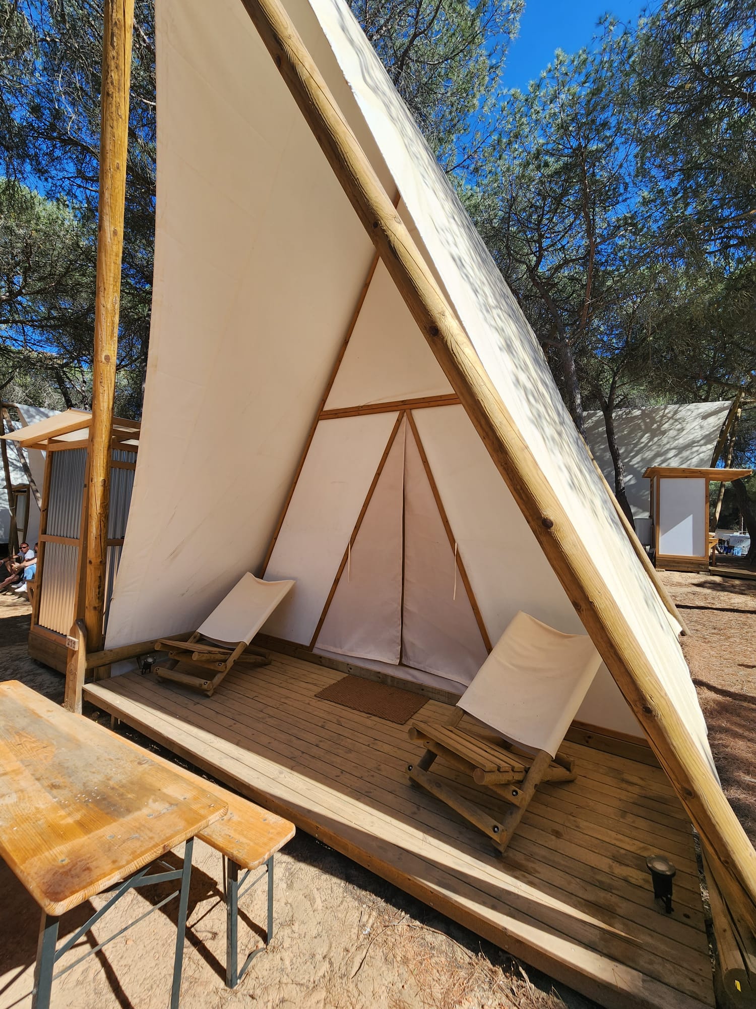 A triangular canvas tent with a wooden frame stands in a forested area, featuring two reclining canvas chairs on a wooden deck. A wooden table is positioned in the foreground, and tall trees are visible under a clear blue sky.