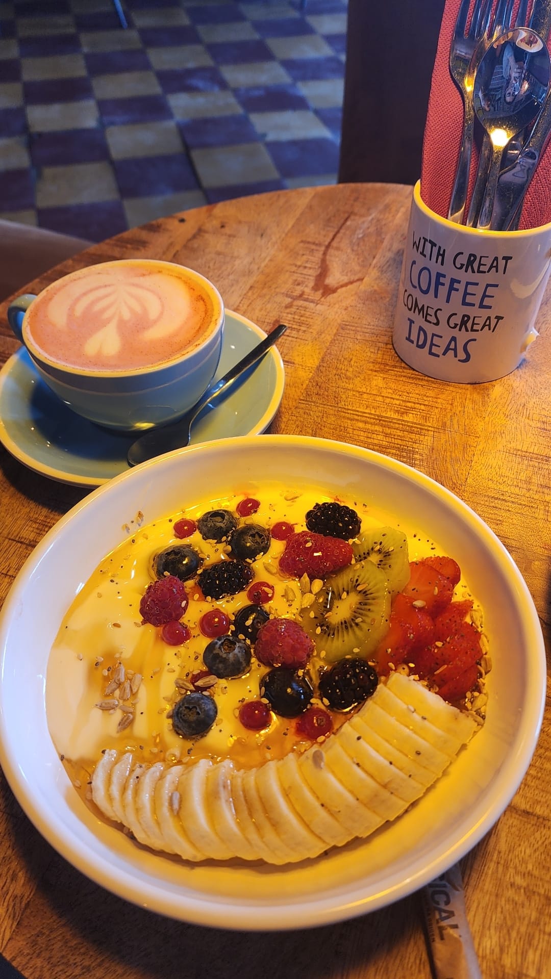 A vibrant breakfast scene features a bowl of yogurt topped with sliced banana, kiwi, strawberries, blueberries, blackberries, and raspberries, sprinkled with seeds. Beside it, a cup of latte with latte art sits on a saucer, and a mug holding cutlery with the phrase "With great coffee comes great ideas" is nearby on the wooden table.