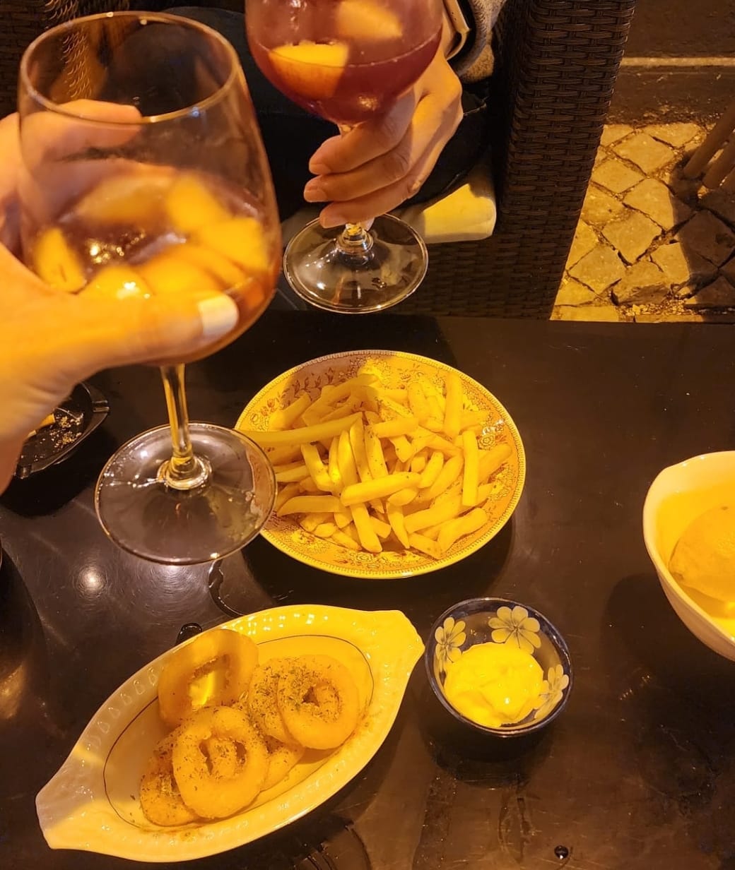 Two people clink glasses filled with a fruity drink, while a table is set with golden fries, crispy onion rings, and a small bowl of yellow dipping sauce, creating a cozy outdoor dining atmosphere.
