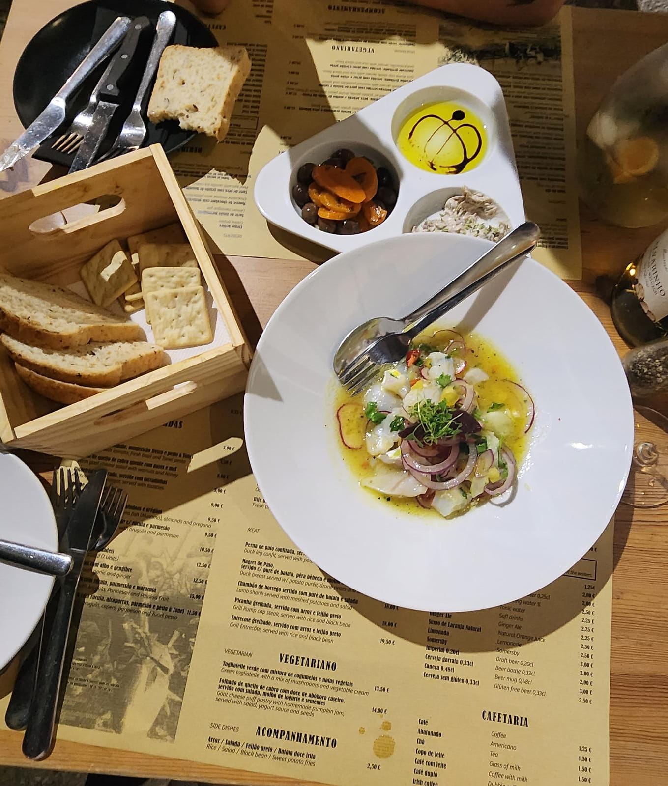A wooden table set with a vibrant dish of ceviche garnished with red onion and herbs, surrounded by a trio of small bowls containing olive oil with balsamic vinegar, olives with apricots, and a creamy spread. A wooden box holds slices of bread and crackers, while utensils rest on a black plate nearby. A menu is partially visible beneath the dishes, adding to the cozy dining atmosphere.