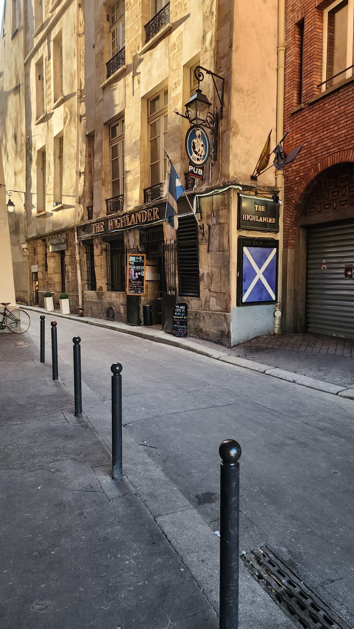 A quiet street scene features "The Highlander" pub, housed in a historic stone building with large windows. A Scottish flag and pub sign hang above the entrance, while chalkboards display menu specials. Black bollards line the sidewalk, and a bicycle is parked nearby.