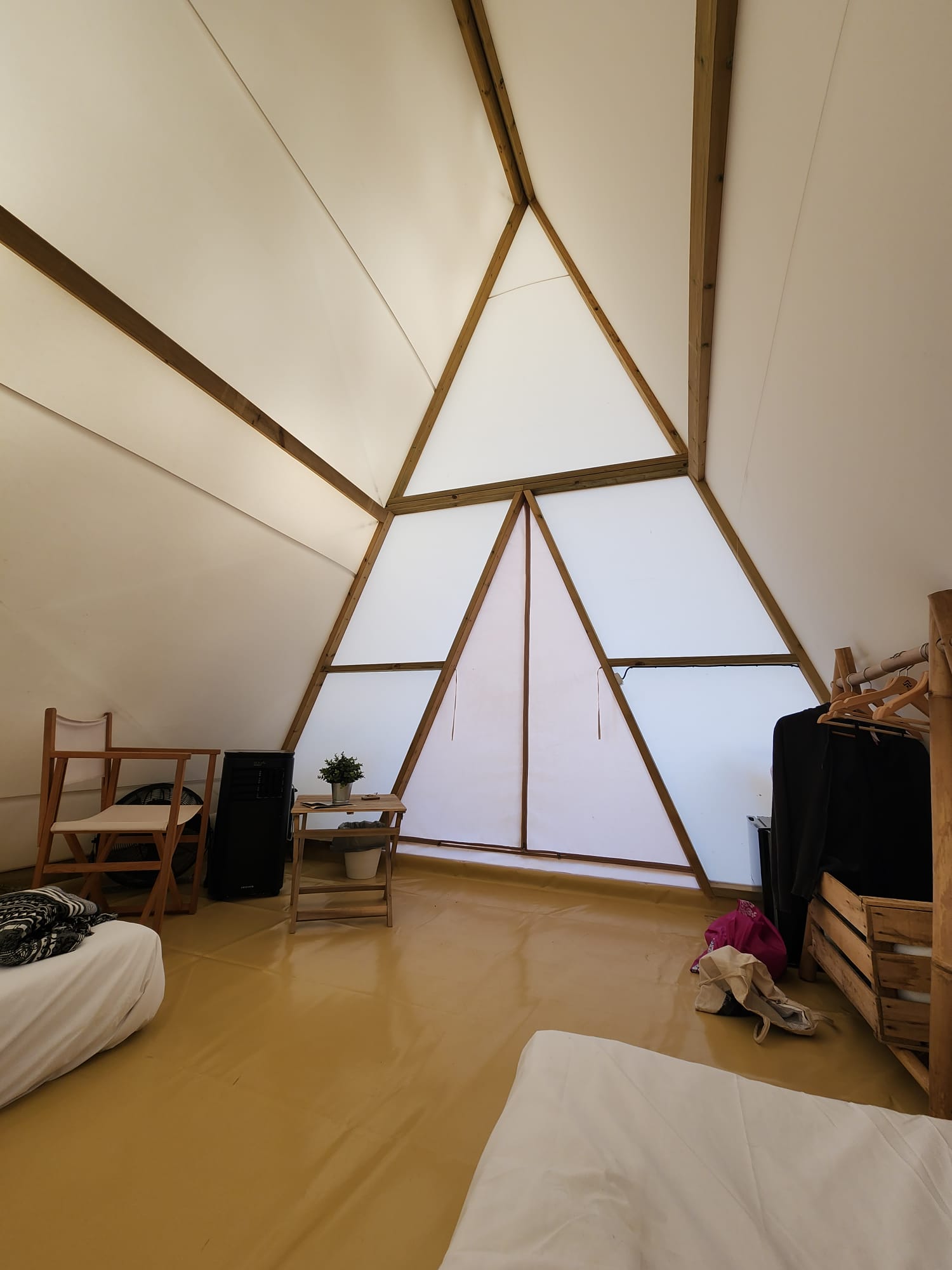 The interior of a triangular tent features two beds with white linens, a wooden chair, a small table with a potted plant, and a clothes rack. The tent has a light-colored floor and wooden frame, creating a cozy, minimalist atmosphere.