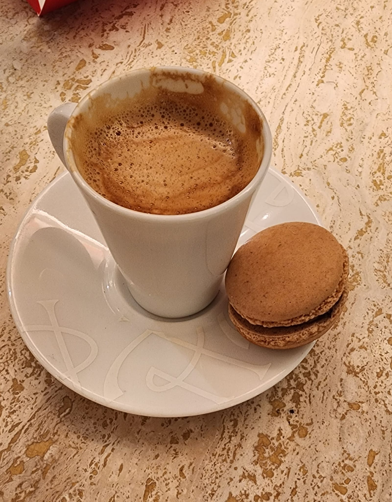 A white cup filled with frothy coffee sits on a matching saucer, accompanied by a single brown macaron, all placed on a textured marble table.