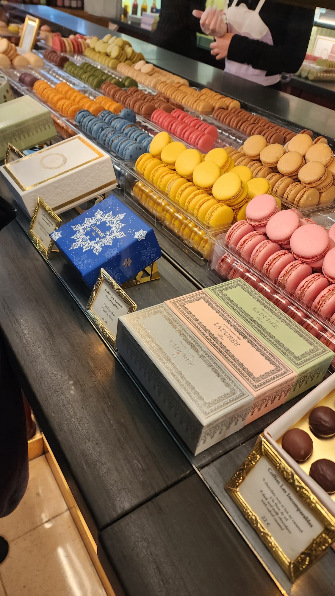 A colorful display of assorted macarons in a patisserie, featuring rows of yellow, pink, blue, and brown macarons. Elegant boxes with intricate designs are arranged in front, and a person in an apron is visible in the background.
