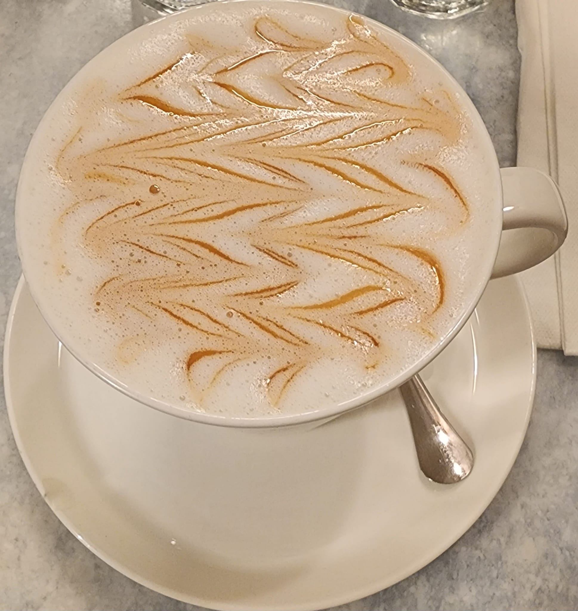 A cup of coffee with creamy foam topped with an intricate caramel drizzle design sits on a white saucer, accompanied by a silver spoon, on a marble tabletop.