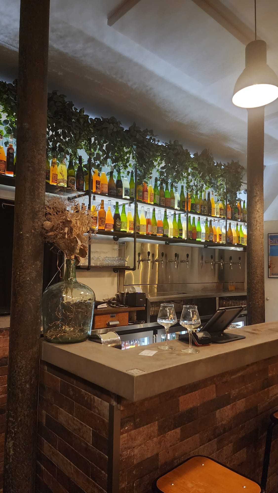 A cozy bar interior featuring a rustic brick counter with two empty wine glasses. Behind the counter, a row of colorful bottles is displayed on shelves, adorned with green foliage. Below the shelves, several taps are visible. A large glass vase with dried flowers sits on the counter, creating a warm and inviting atmosphere.