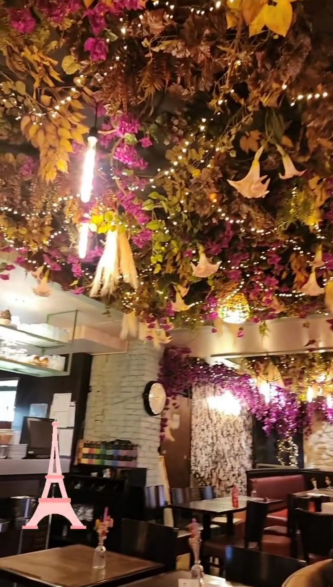 A cozy café interior with a ceiling adorned with vibrant purple and green foliage and twinkling string lights. Tables are neatly arranged below, each with a small vase of flowers, creating a warm and inviting atmosphere. A pink Eiffel Tower icon is visible in the foreground.
