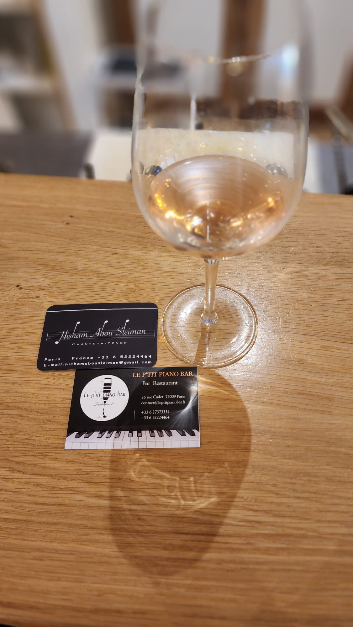 A glass of white wine sits on a wooden table, accompanied by two business cards. The cards display contact information and branding for a singer and a piano bar, adding a touch of sophistication to the scene.
