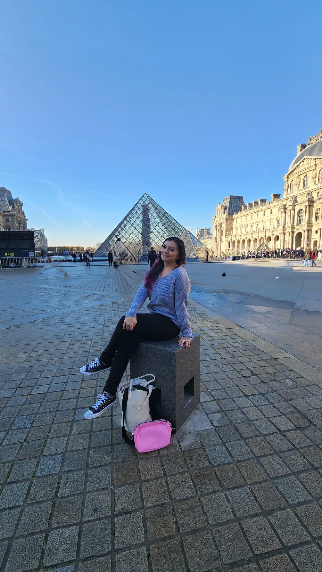 A person sits on a stone block in front of the Louvre Pyramid, wearing a purple sweater and black pants. A white tote bag and a pink pouch rest on the ground nearby. The clear blue sky and surrounding historic architecture create a picturesque scene.