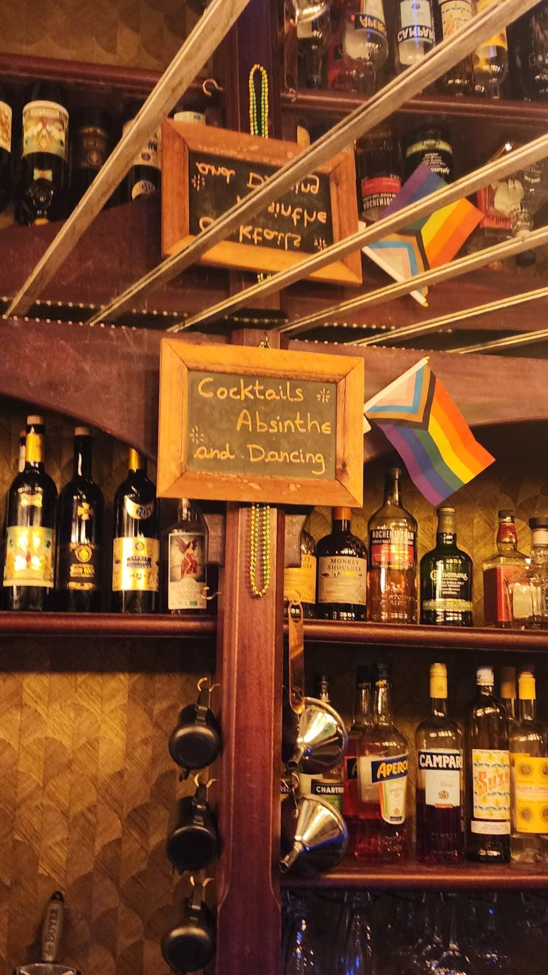 A cozy bar shelf displays various liquor bottles, including Aperol and Campari, with a small pride flag nearby. A chalkboard sign reads "Cocktails, Absinthe, and Dancing," adding to the inviting atmosphere.