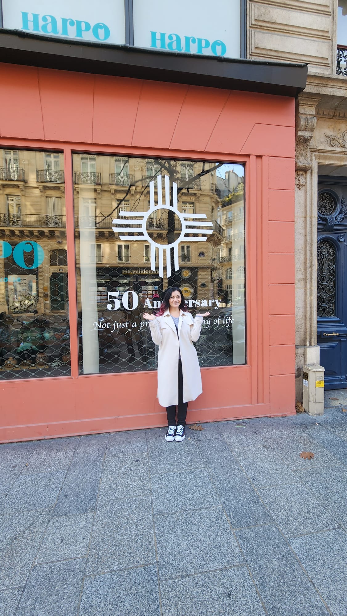 A woman in a light coat and sneakers stands smiling in front of a shop with a large window display celebrating a 50th anniversary. The building features a distinctive orange facade with reflections of nearby architecture in the glass.