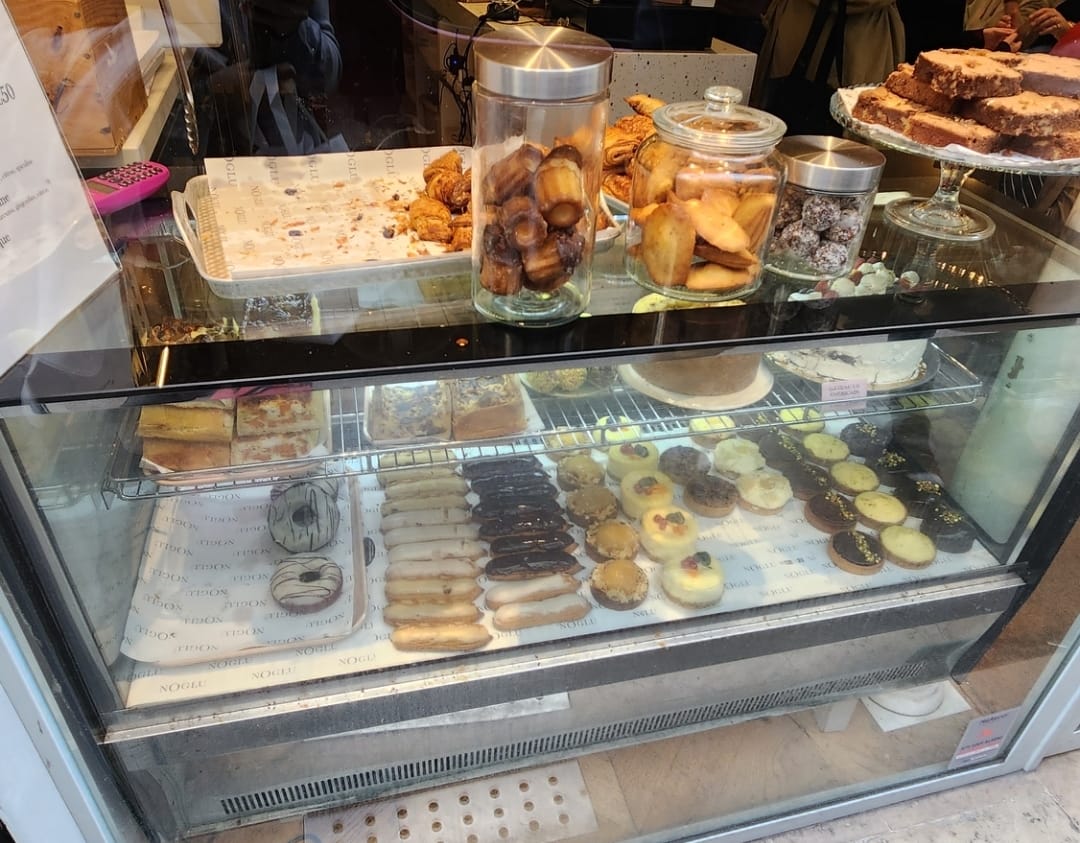 A bakery display case filled with an assortment of pastries and desserts. The top shelf features glass jars of cookies and pastries, along with a tray of croissants. Below, there are rows of eclairs, tarts, and other sweet treats, all neatly arranged on trays. The setting is cozy and inviting, showcasing a variety of tempting baked goods.