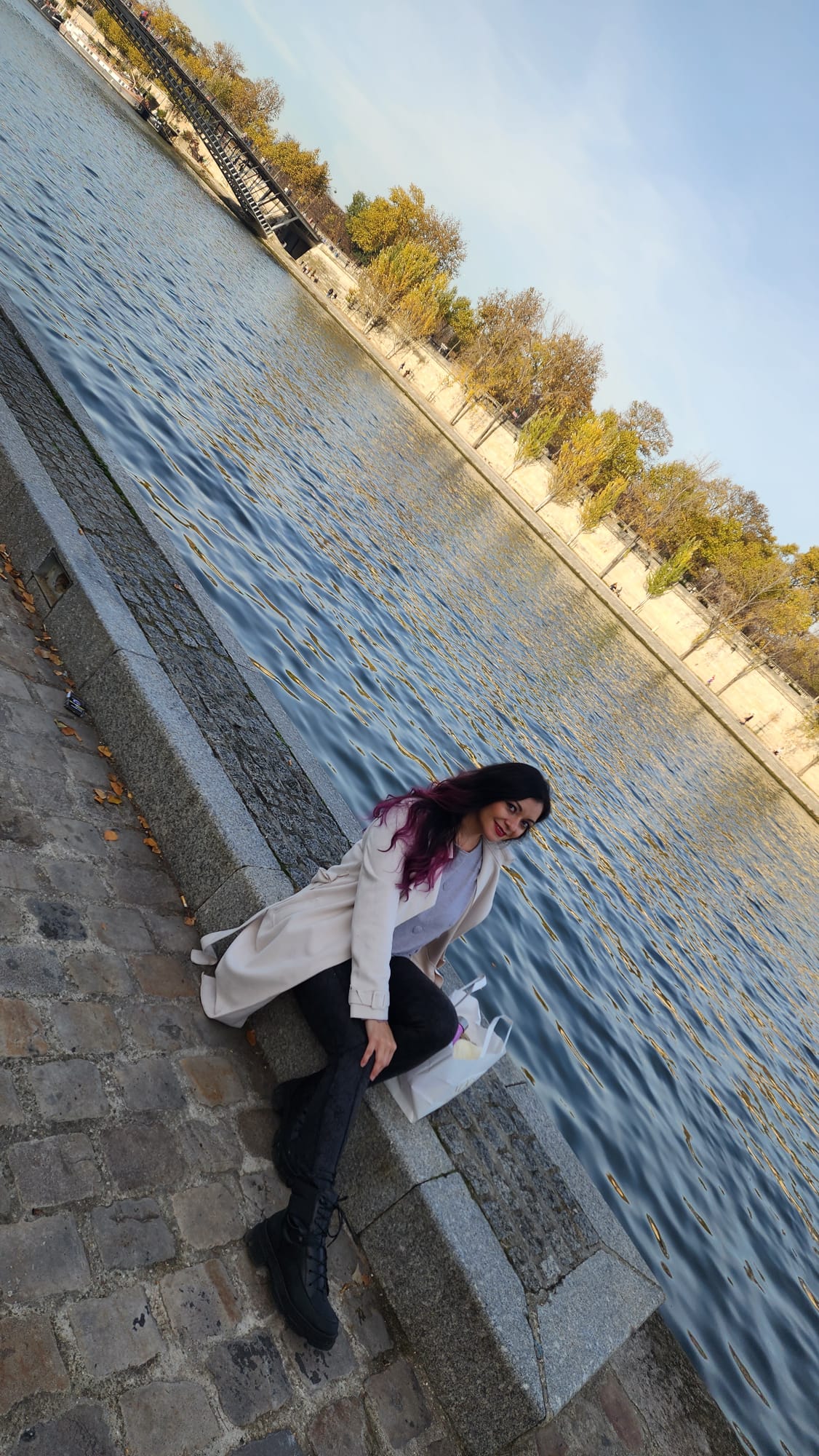 A person with long, dark hair sits on a stone ledge by a river, smiling. They wear a light coat and black boots, with a shopping bag beside them. The background features a tree-lined riverbank under a clear blue sky.