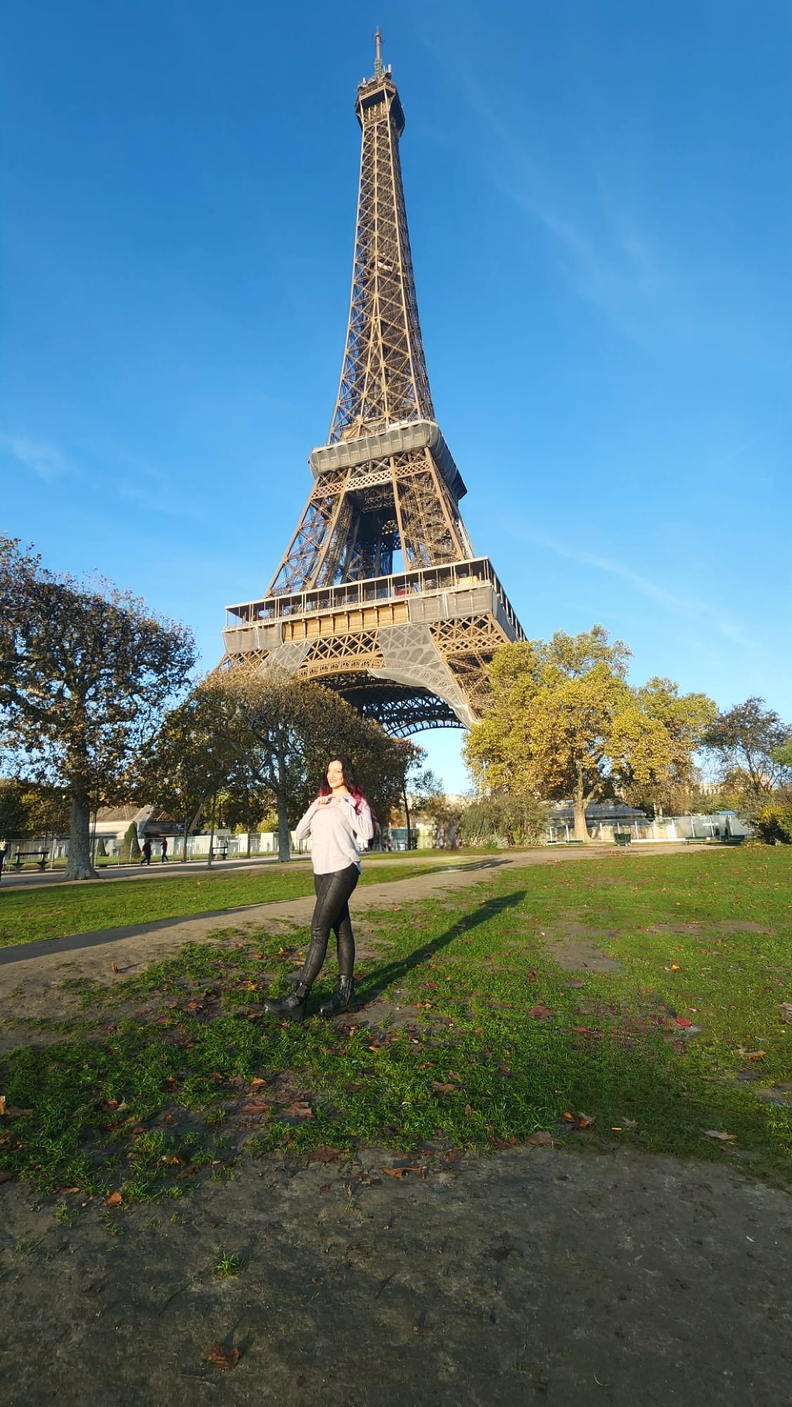 A person stands in front of the Eiffel Tower on a sunny day, posing on a grassy area with scattered autumn leaves, surrounded by trees with some green and golden foliage.