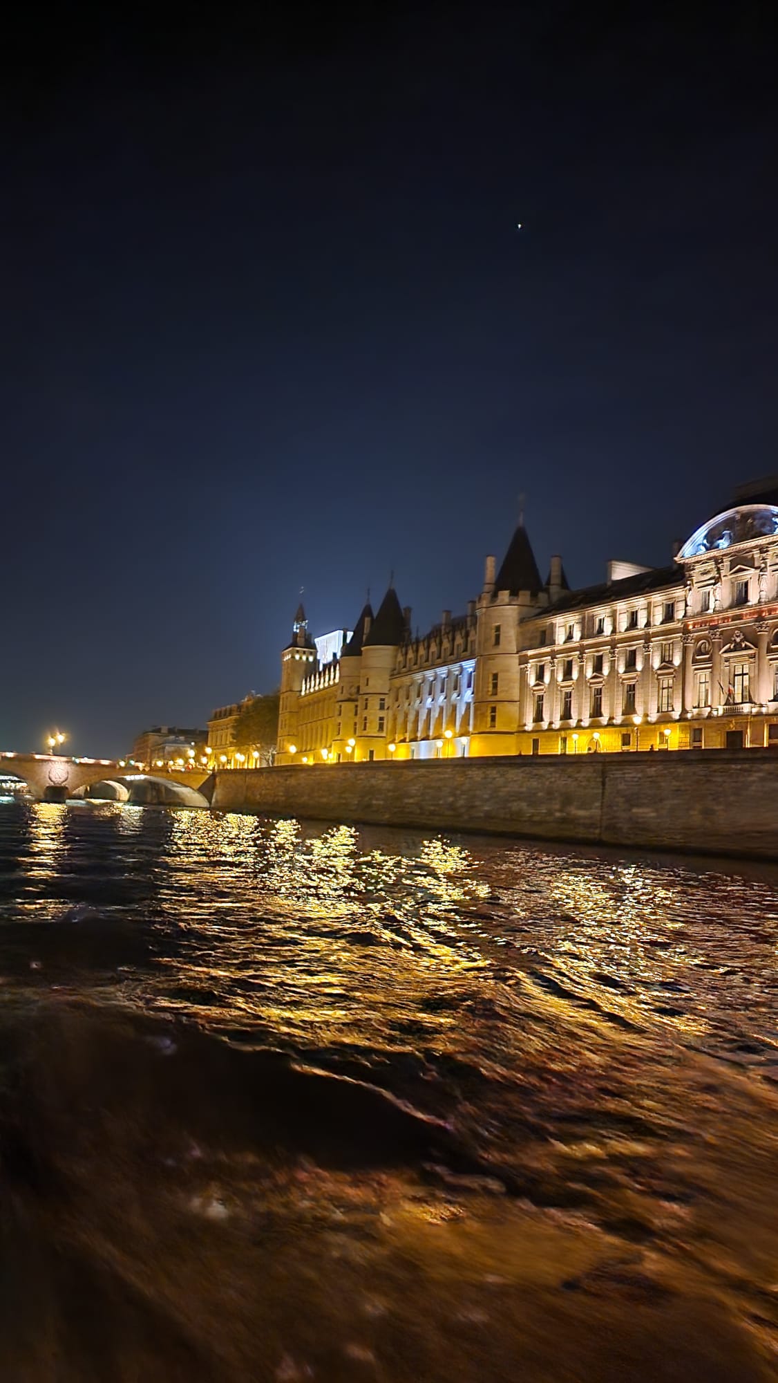 A beautifully illuminated riverside building at night, with its lights reflecting on the gently rippling water. A bridge in the background is also lit, adding to the serene nighttime scene under a clear, dark sky.
