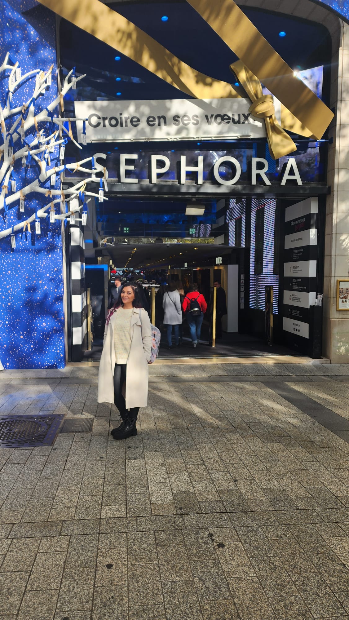 A person in a light coat stands smiling in front of a Sephora store entrance, adorned with festive blue decorations and a large gold ribbon. The store is bustling with people entering and exiting.