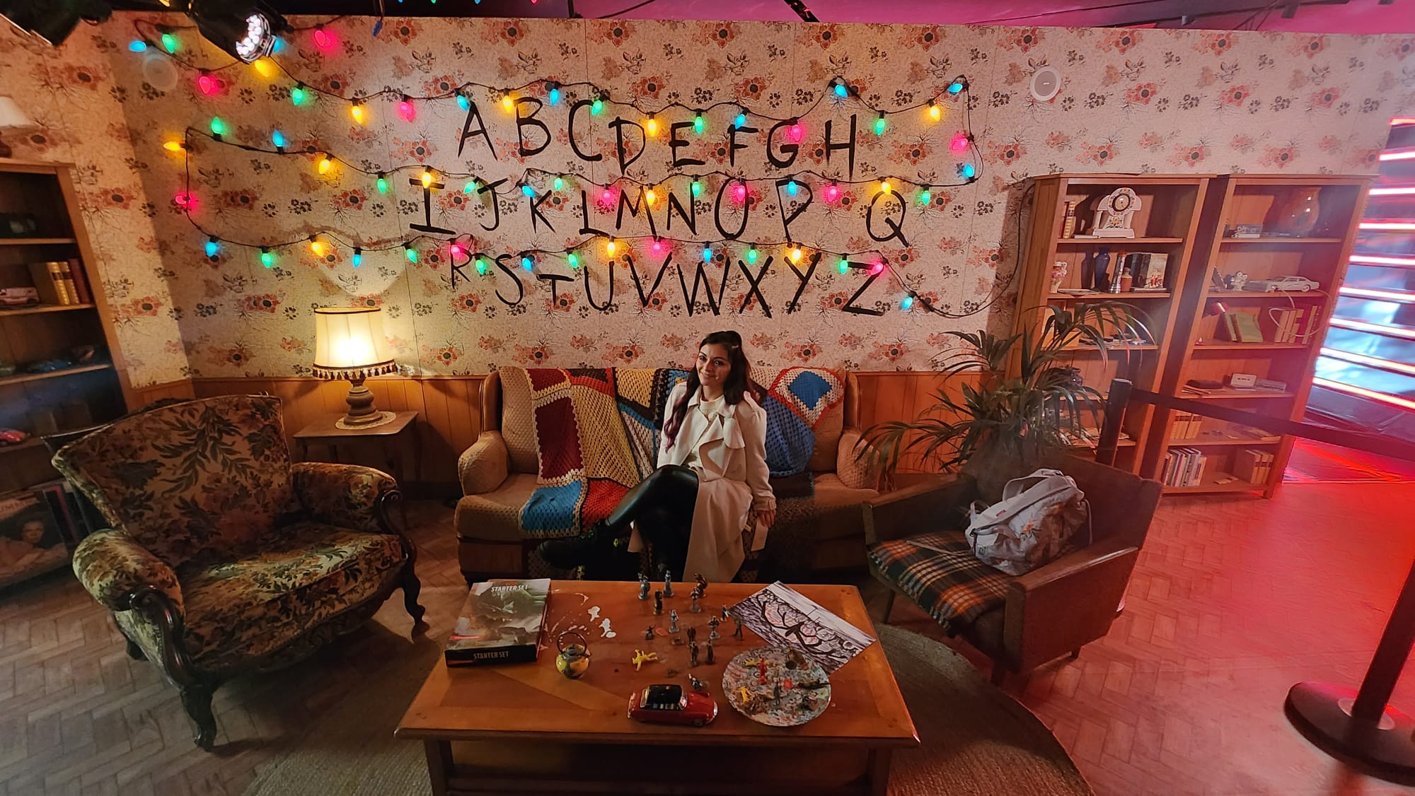 A woman sits on a vintage couch in a cozy room with floral wallpaper. The wall behind her features the alphabet written in black, adorned with colorful string lights. The room is warmly lit, with a patterned armchair, a wooden coffee table displaying figurines, and bookshelves filled with various items.