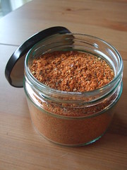 A glass jar filled with a reddish-brown spice mix sits on a wooden table, with its black lid resting nearby.
