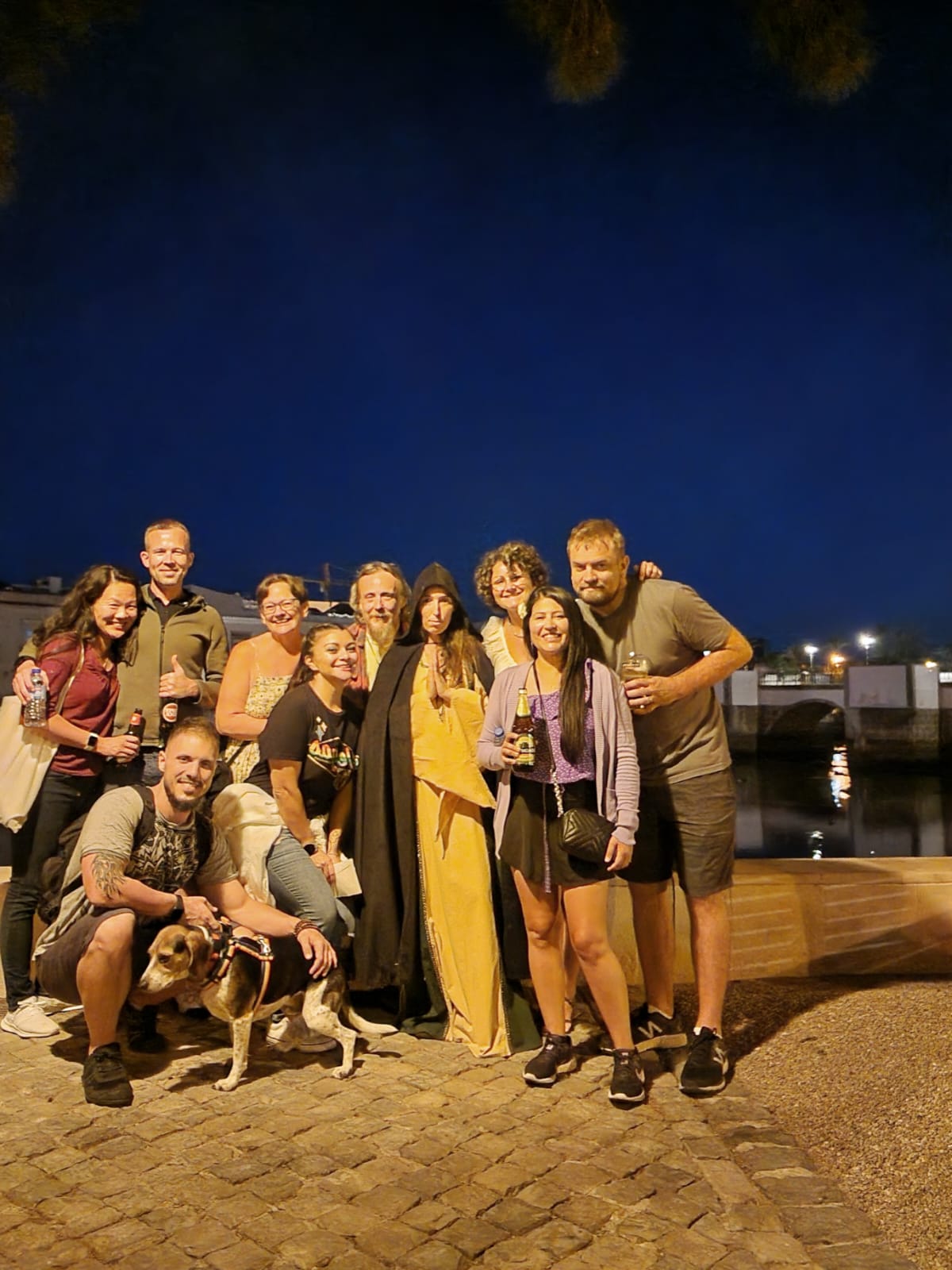 A group of people gathers on a cobblestone street at night, posing for a photo. Some hold drinks, while one person is dressed in a hooded robe. A man crouches in front, smiling with a dog on a leash. The background features a river and a bridge under a dark, clear sky.