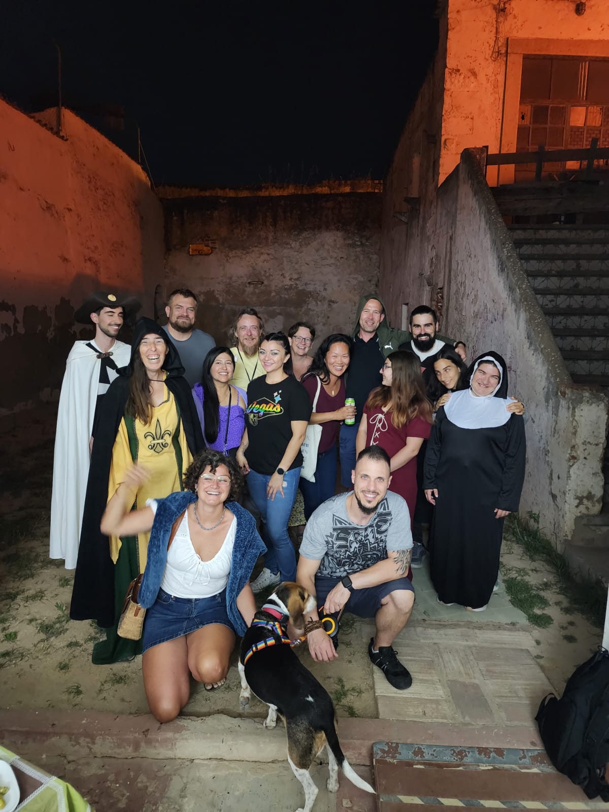 A group of people, some in costumes, pose for a photo in an outdoor setting with a rustic wall and stairs in the background. A woman kneels in front, smiling, next to a dog wearing a colorful harness. The atmosphere is cheerful and casual.