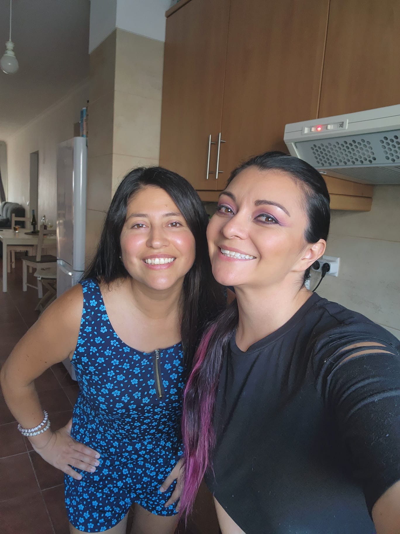 Two smiling women pose for a selfie in a kitchen. One wears a blue floral outfit, while the other sports a black top with purple accents in her hair. The kitchen features wooden cabinets and a visible range hood.