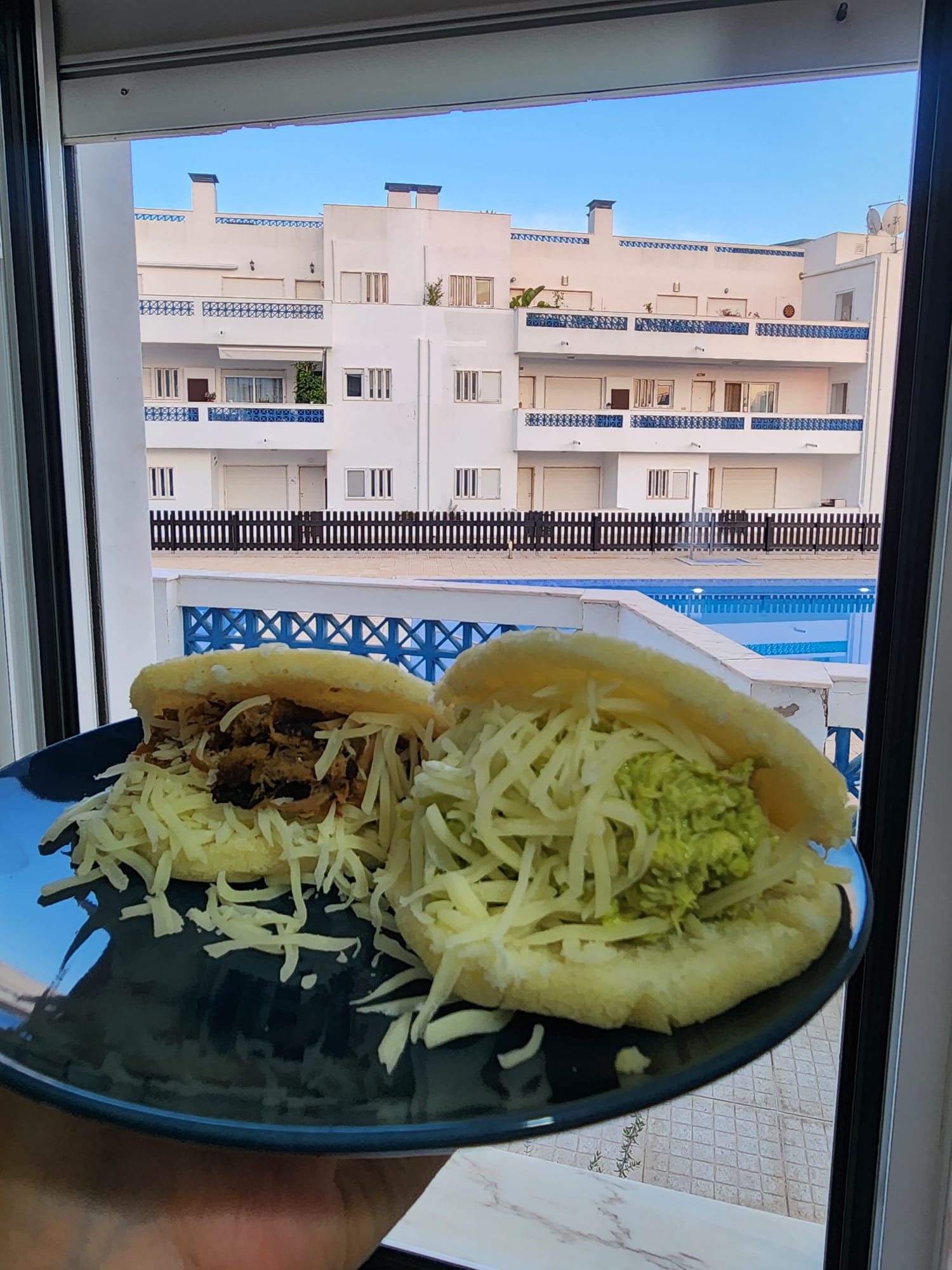 A plate holds two arepas filled with shredded cheese, one with pulled meat and the other with guacamole, against a backdrop of white residential buildings and a blue sky seen through a window.