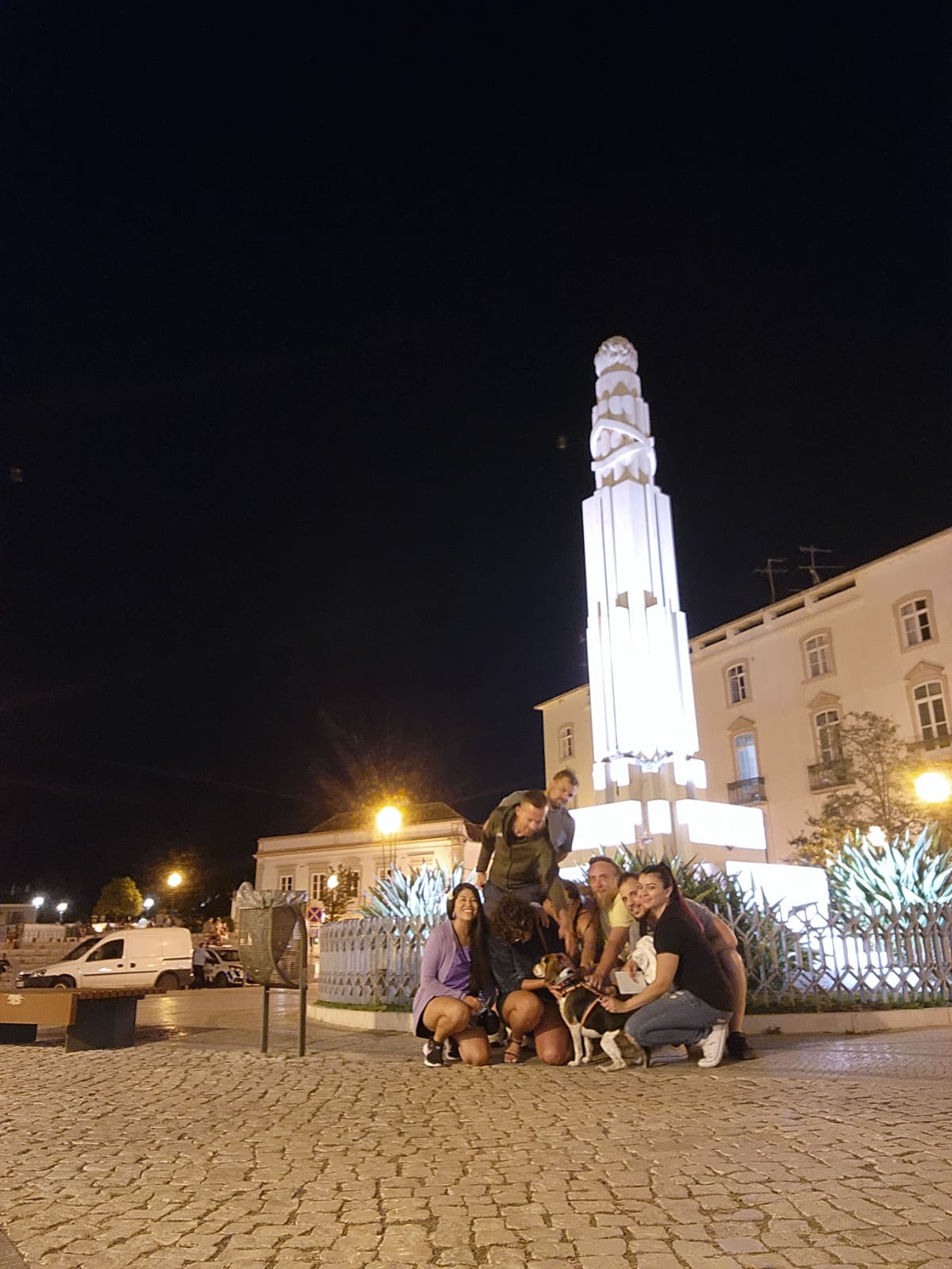 A group of people and a dog gather on a cobblestone square at night, with a tall, illuminated white monument in the background. The scene is warmly lit by streetlights, and a few parked vehicles are visible in the distance.