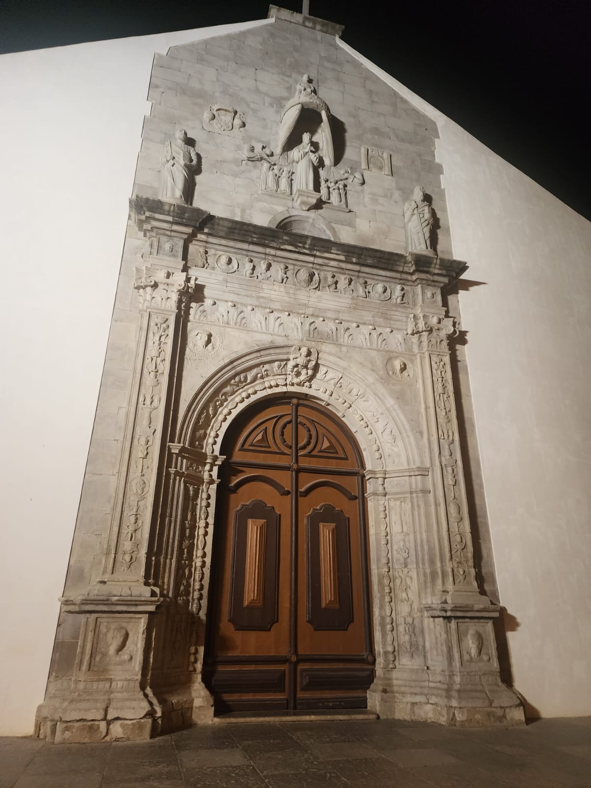 A grand, ornately carved stone doorway with a wooden door stands illuminated at night. The facade features intricate sculptures and reliefs, including figures and decorative elements, set against a smooth, white wall.