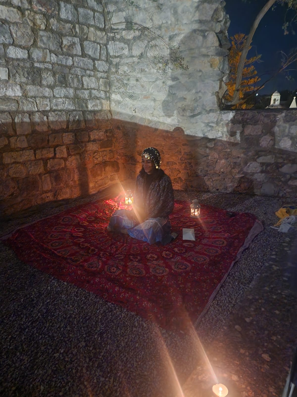 A person sits cross-legged on a patterned red rug, surrounded by lit candles, inside a stone-walled room at night. They wear a dark outfit with a headpiece, creating a serene and mystical atmosphere.