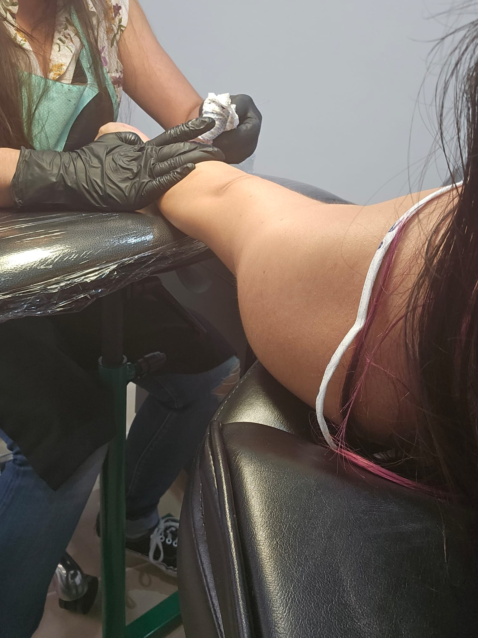 A tattoo artist wearing black gloves is working on a person's arm, which is resting on a padded chair. The artist is using a cloth to wipe the area, and the client has long dark hair with a hint of pink.