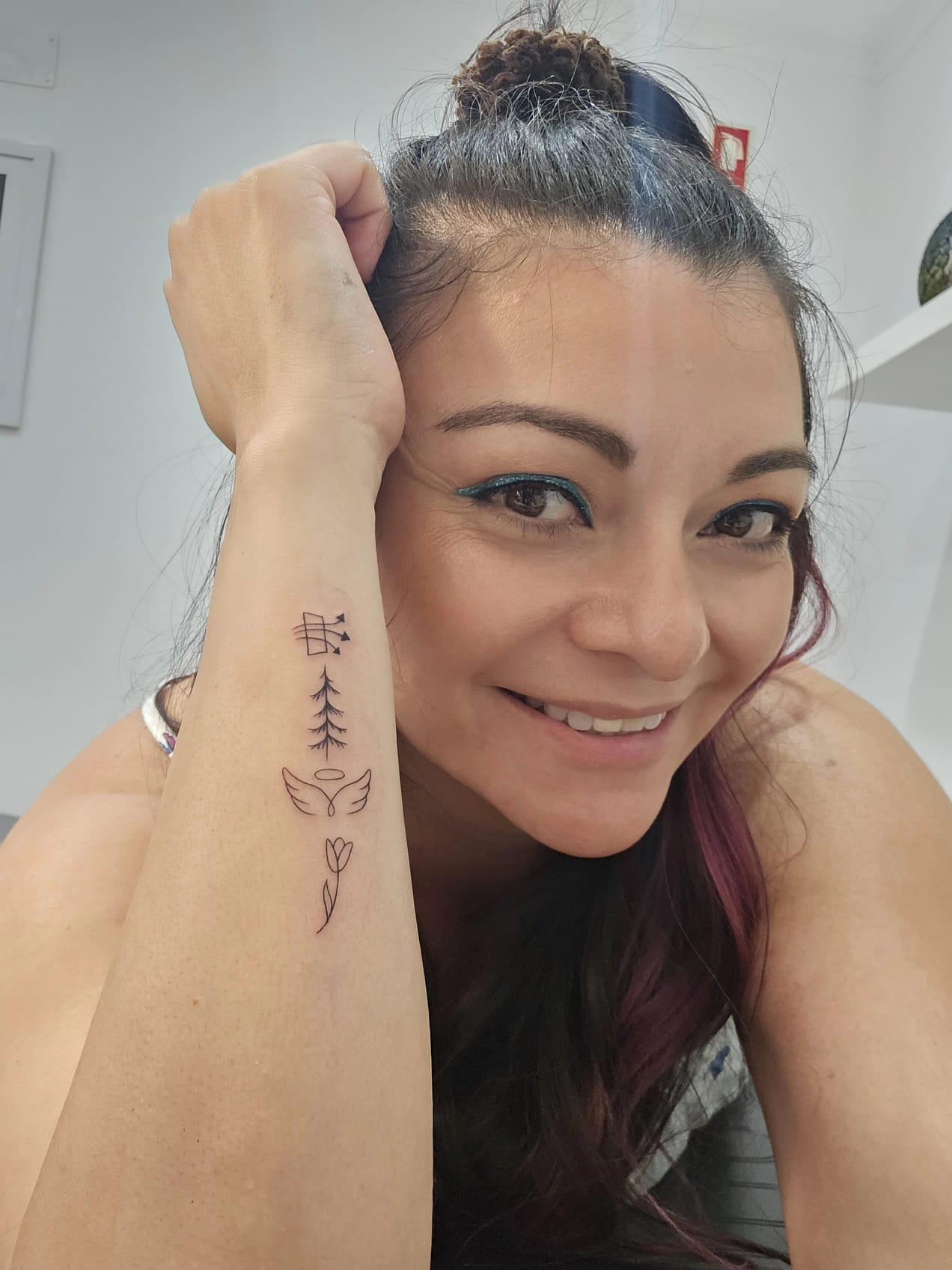 A woman with a top bun smiles while showing a forearm tattoo featuring a series of minimalist symbols including a butterfly, tree, angel wings, and a tulip.