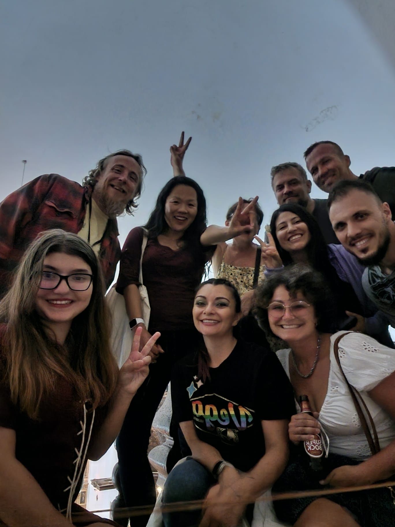 A group of people smiling and making peace signs, gathered closely together. They appear to be in a casual indoor setting, with one person holding a drink bottle. The mood is cheerful and friendly.