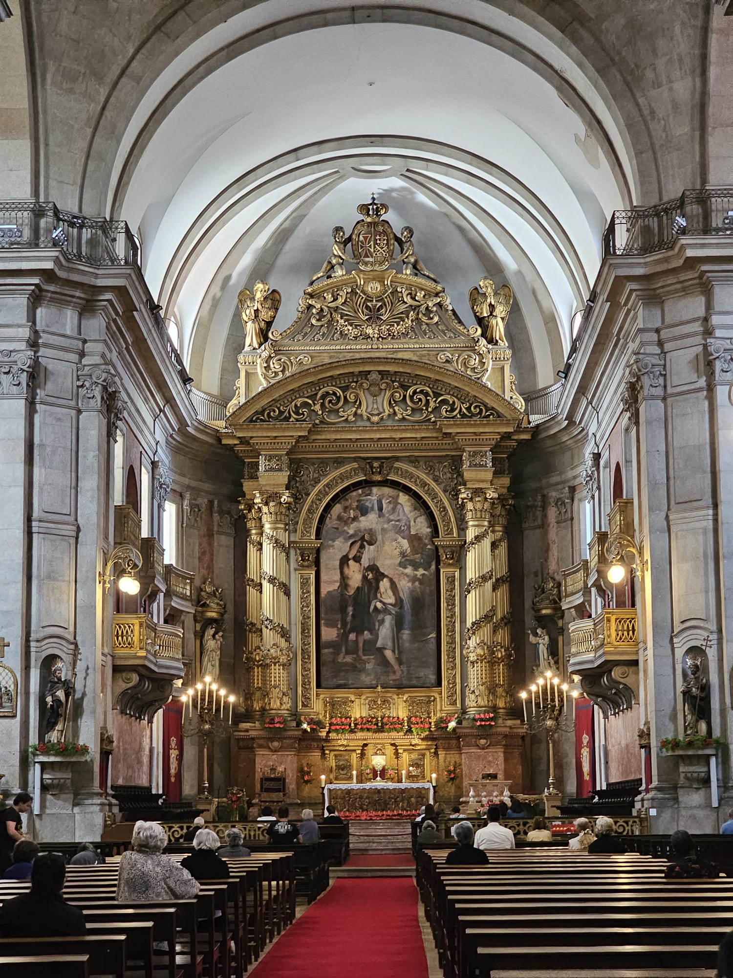 The interior of an ornate church features a grand altar with intricate gold detailing and a large religious painting. The high arched ceiling and stone walls add to the majestic atmosphere. People are seated on wooden pews on a red carpet, facing the altar.