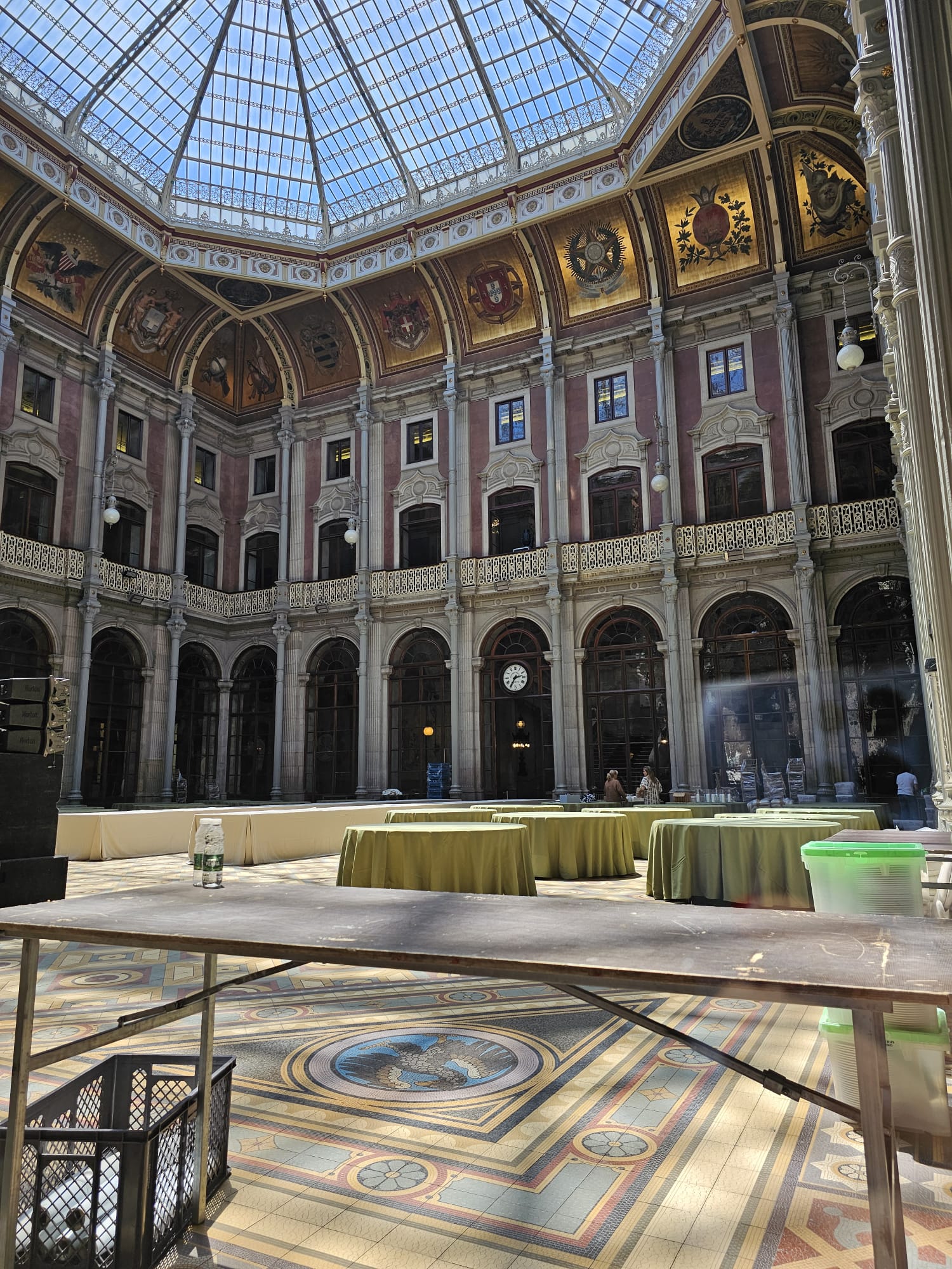 A spacious, ornate atrium with a glass-domed ceiling and elaborate frescoes on the upper walls. The floor features a detailed mosaic, and several round tables with green tablecloths are set up. A clock is visible on the far wall, and natural light floods the elegant space.