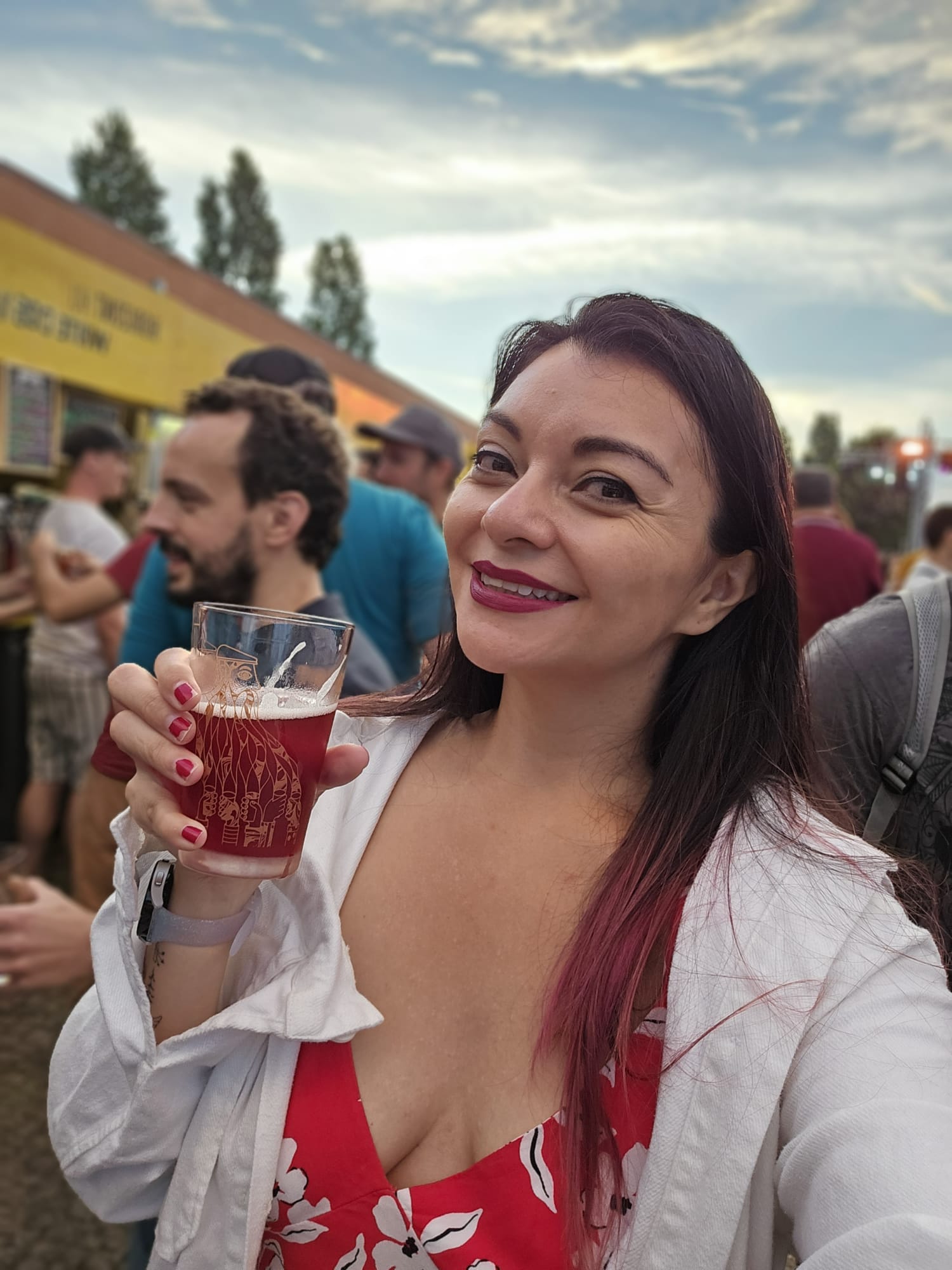 A woman in a red floral dress and white jacket smiles while holding a glass of red beer at an outdoor event. The background shows people mingling near a yellow food stall under a partly cloudy sky.