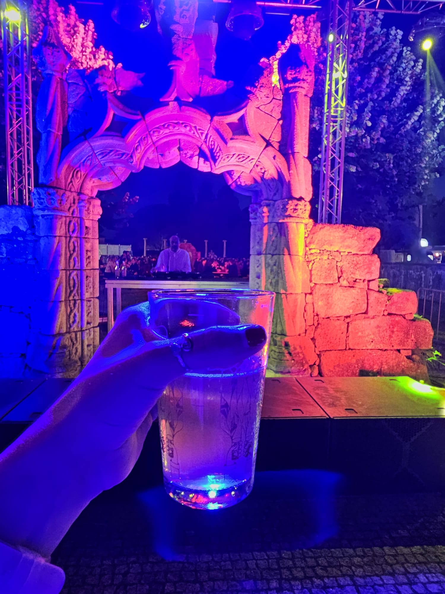 A hand holding a glowing drink in a clear cup, illuminated by colorful lights, with a vibrant, intricately carved stone archway in the background. The scene is set in a lively outdoor atmosphere, with a DJ visible under the arch and colorful lighting creating a festive ambiance.