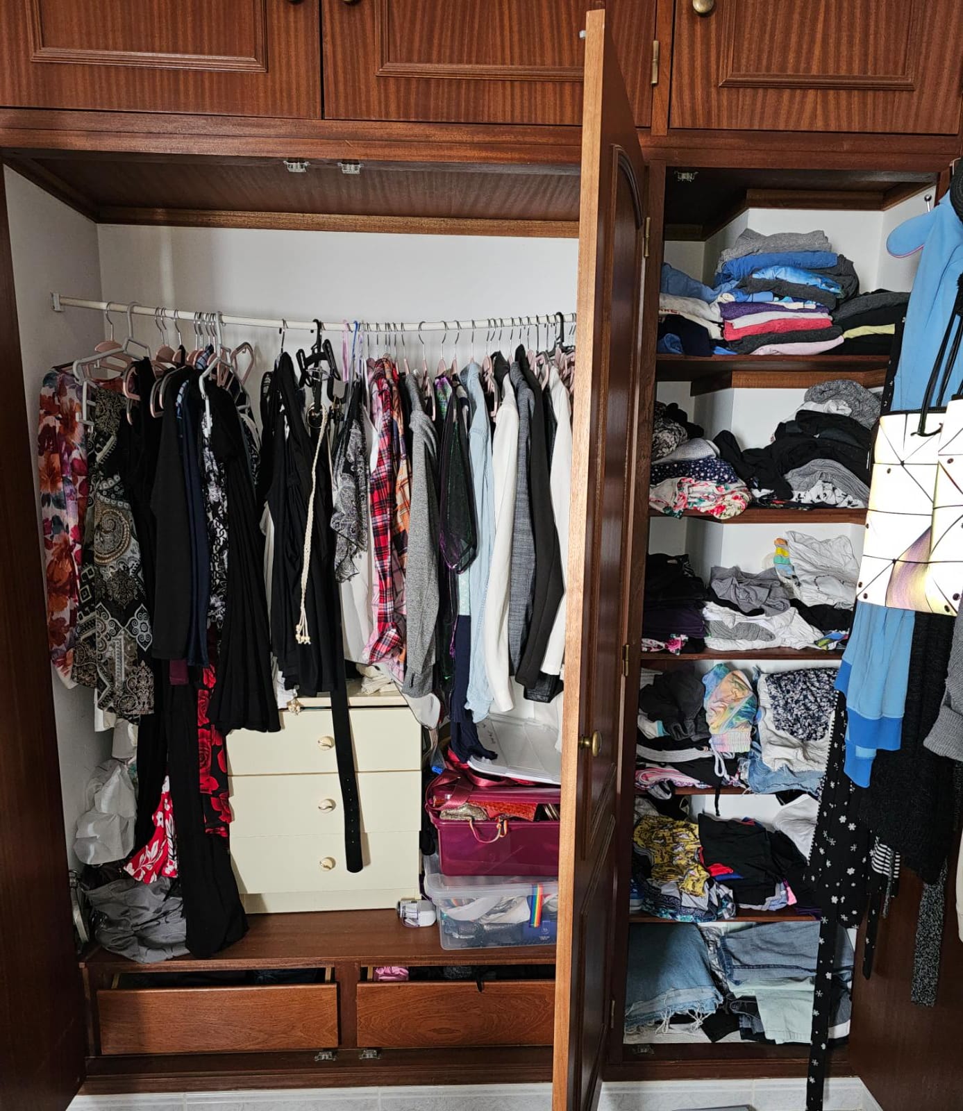 An open wooden wardrobe reveals a collection of clothes on hangers, including patterned and solid tops, and a variety of folded garments on shelves. A small white chest of drawers and plastic storage boxes are visible at the bottom, with some items tucked in the lower drawers.