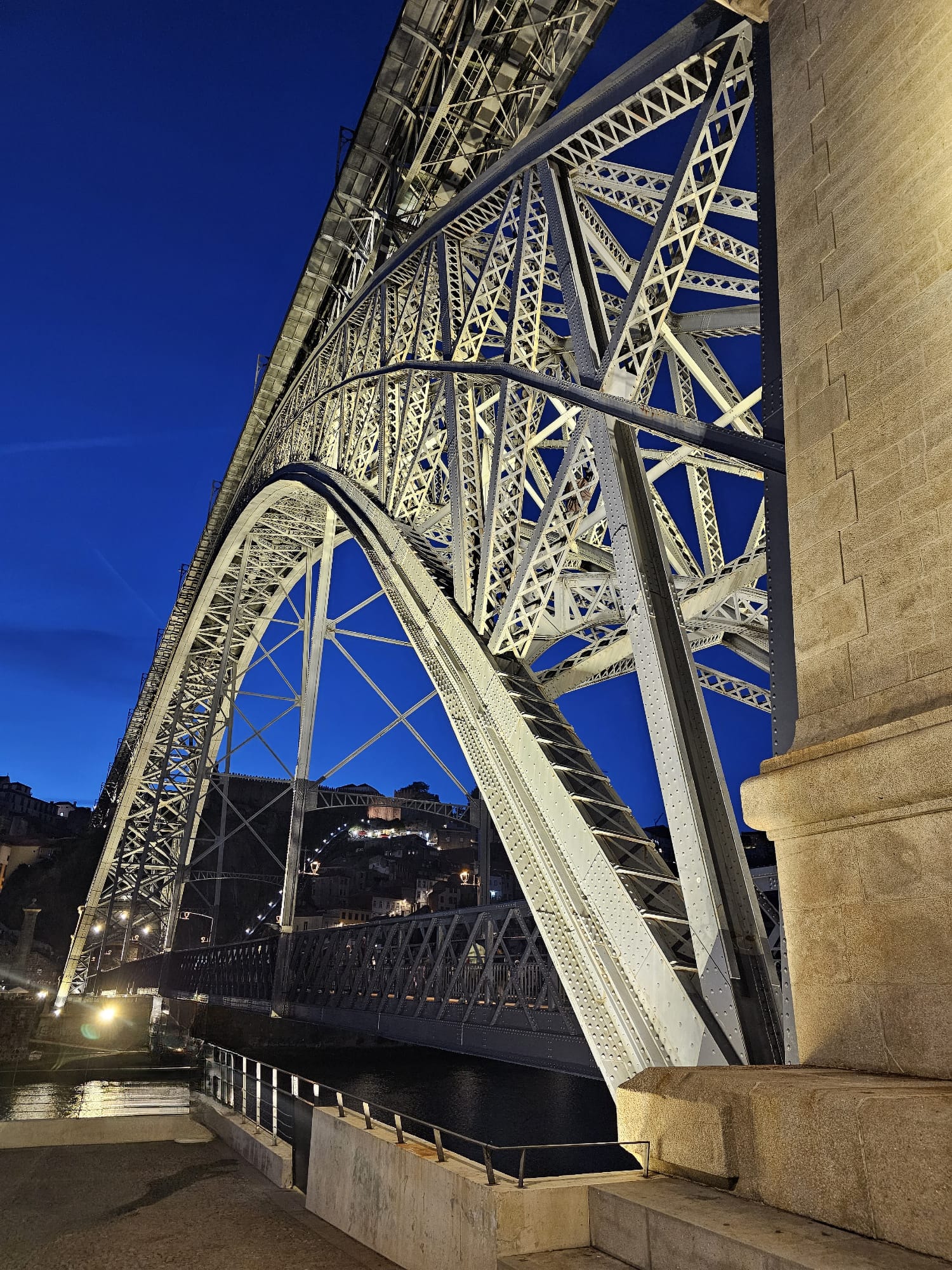 A large, illuminated steel arch bridge spans a river against a deep blue evening sky, with city lights twinkling on the hillside in the background.