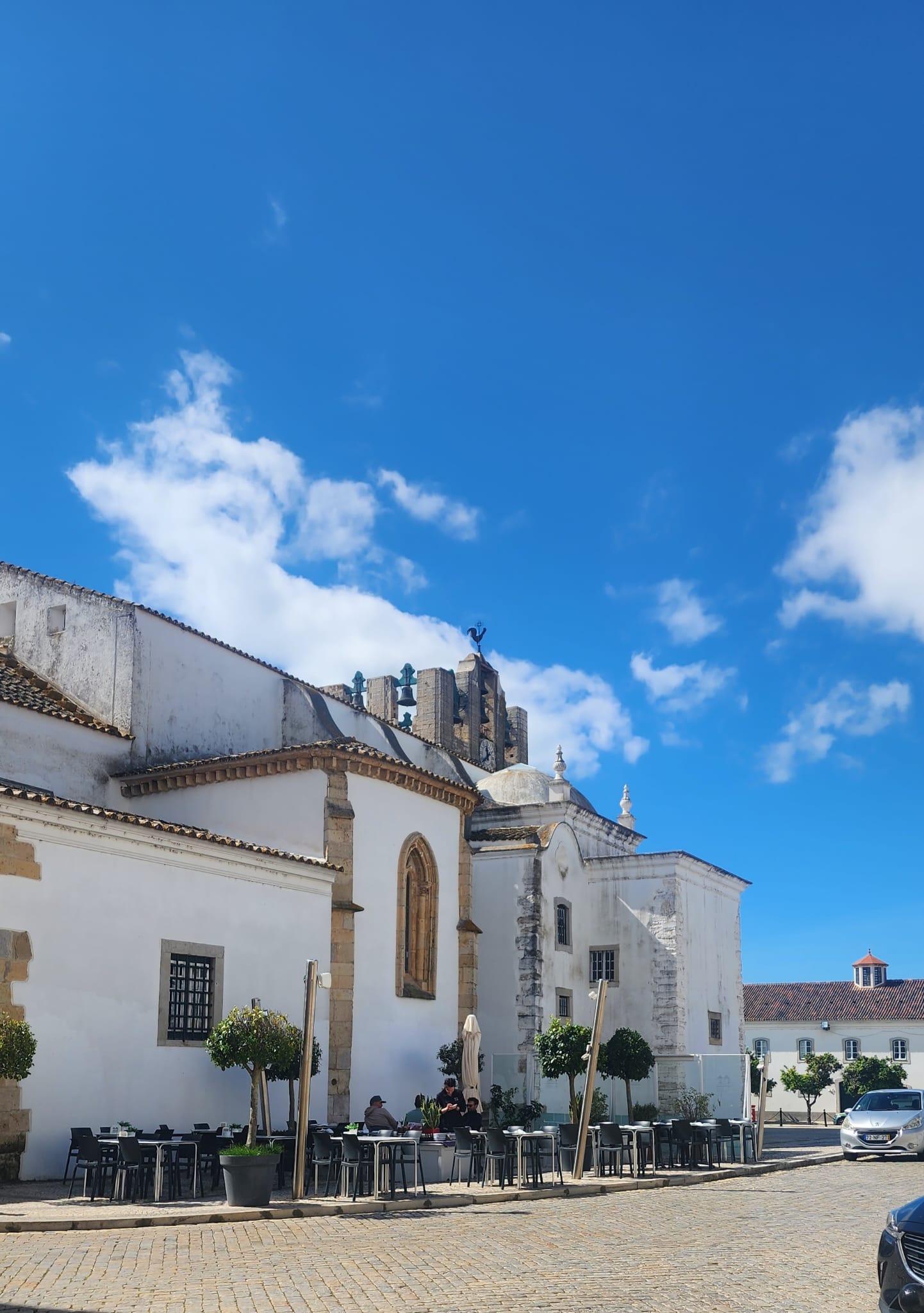 A charming white building with arched windows and a tiled roof stands under a bright blue sky, with a few people seated at outdoor tables surrounded by potted trees on a cobblestone street.