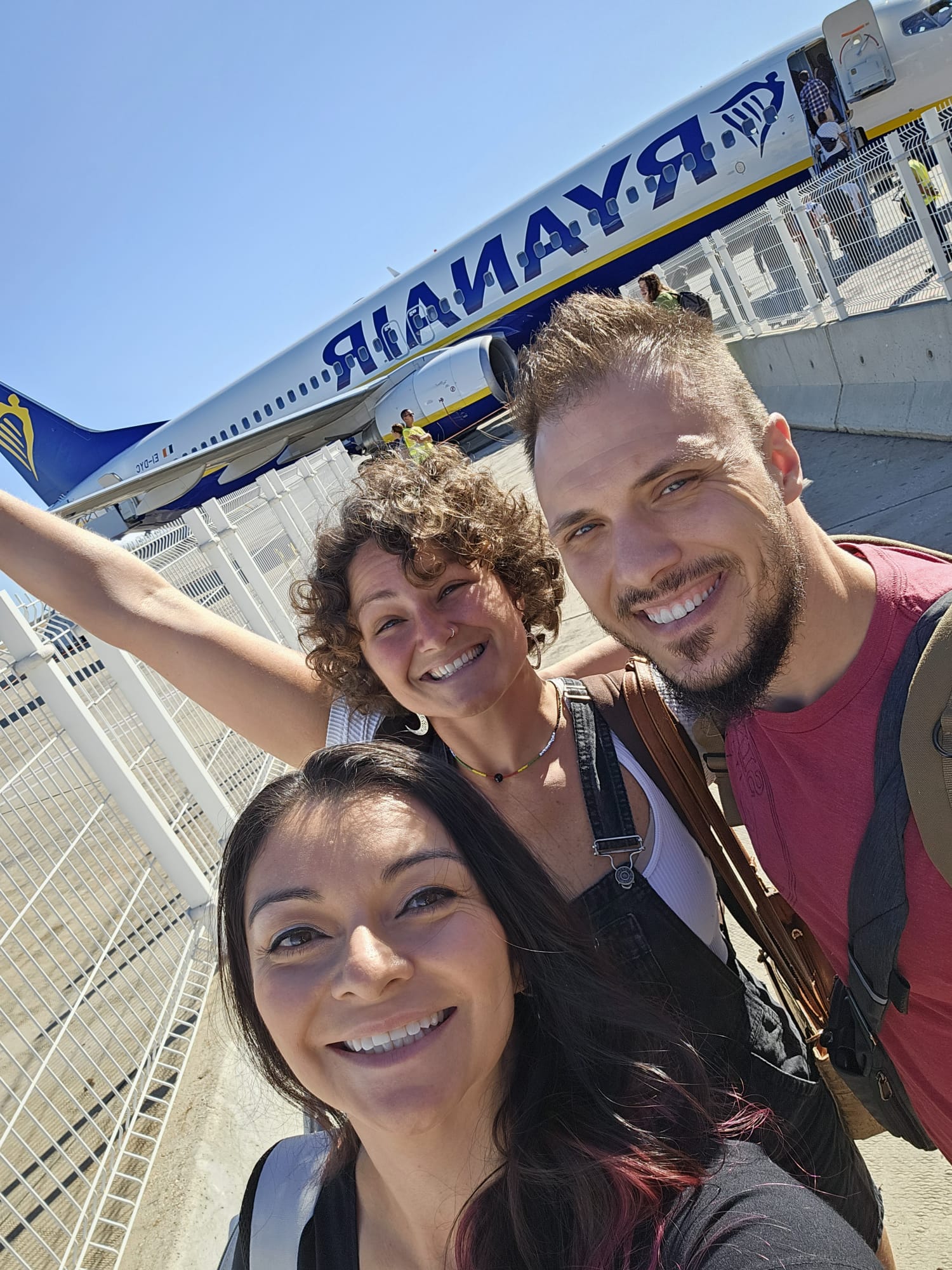 Three people smile for a selfie in front of a Ryanair airplane on a sunny day, with the aircraft visible in the background as passengers board.