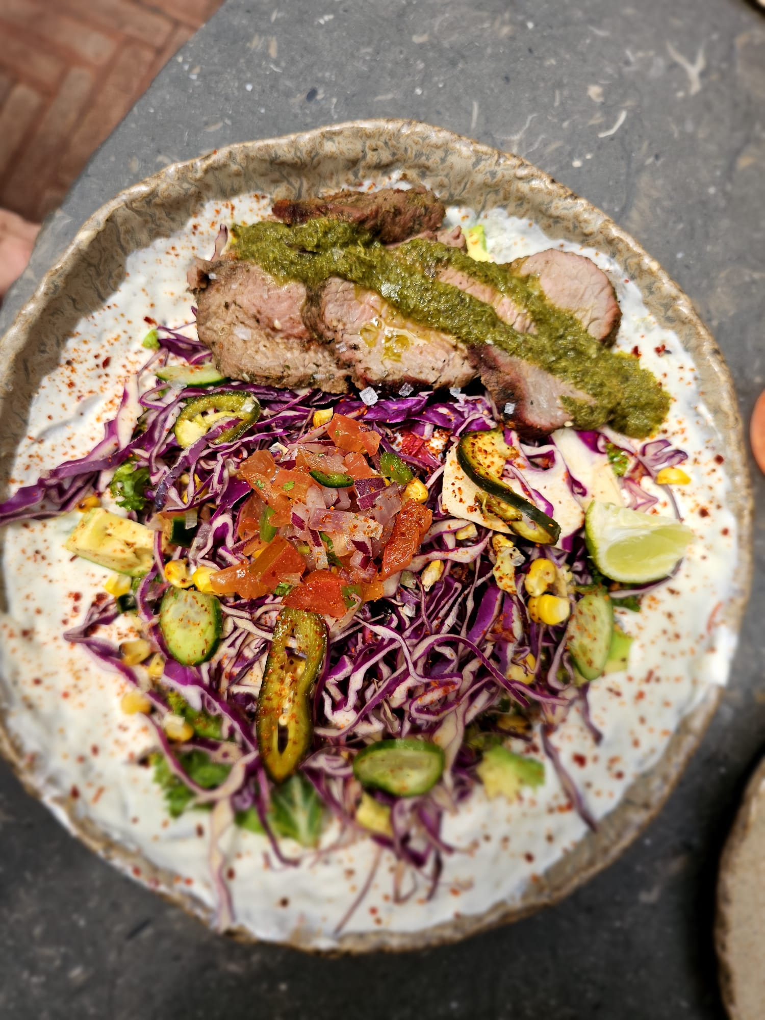 A rustic bowl filled with a vibrant salad topped with slices of grilled meat drizzled with green sauce. The salad features shredded purple cabbage, grilled corn, sliced cucumbers, and a sprinkle of red seasoning, all garnished with a lime wedge and diced tomatoes.