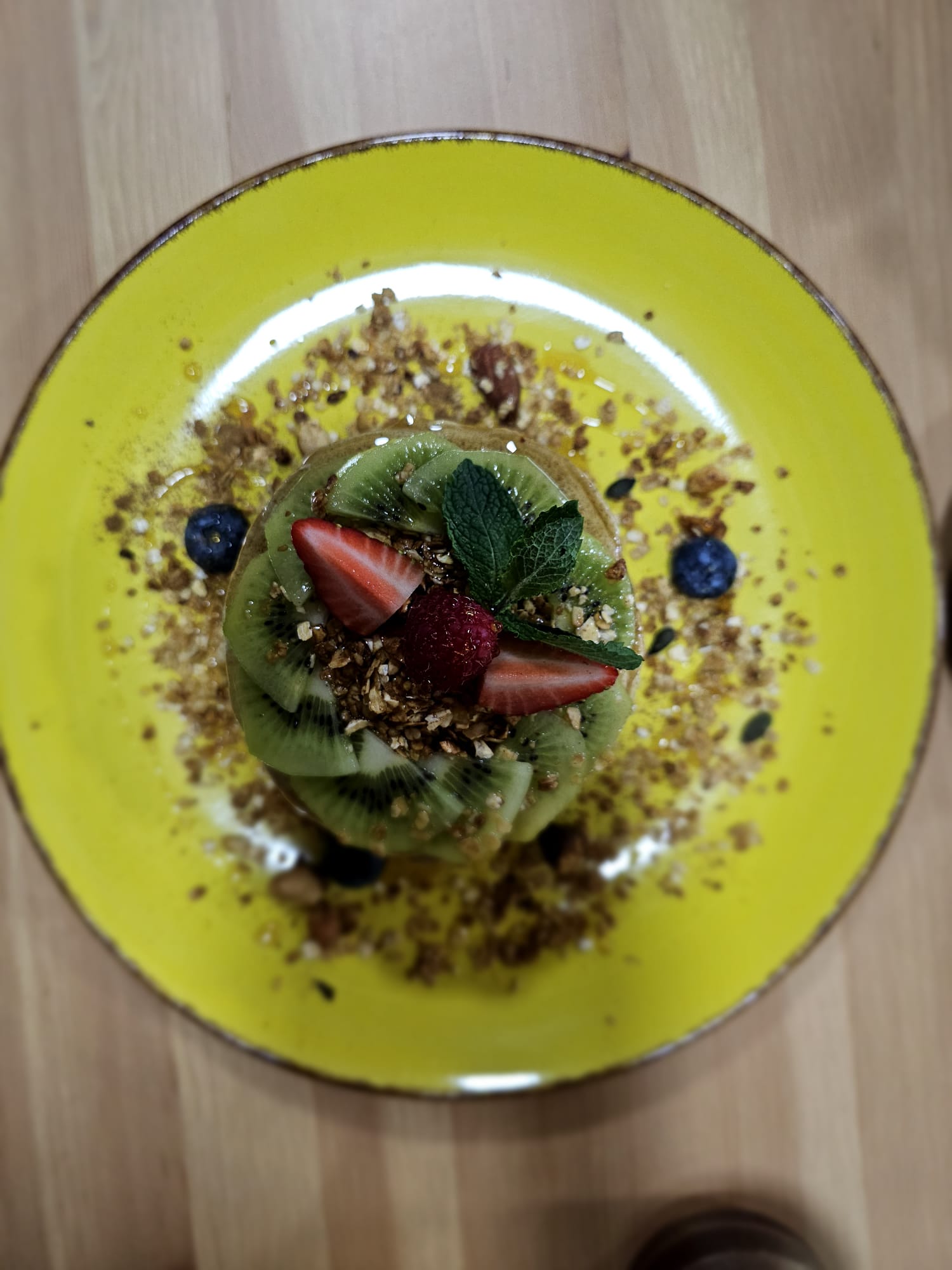 A vibrant dish on a yellow plate features a stack of pancakes topped with sliced kiwi, strawberries, and a sprig of mint, surrounded by scattered granola and blueberries.
