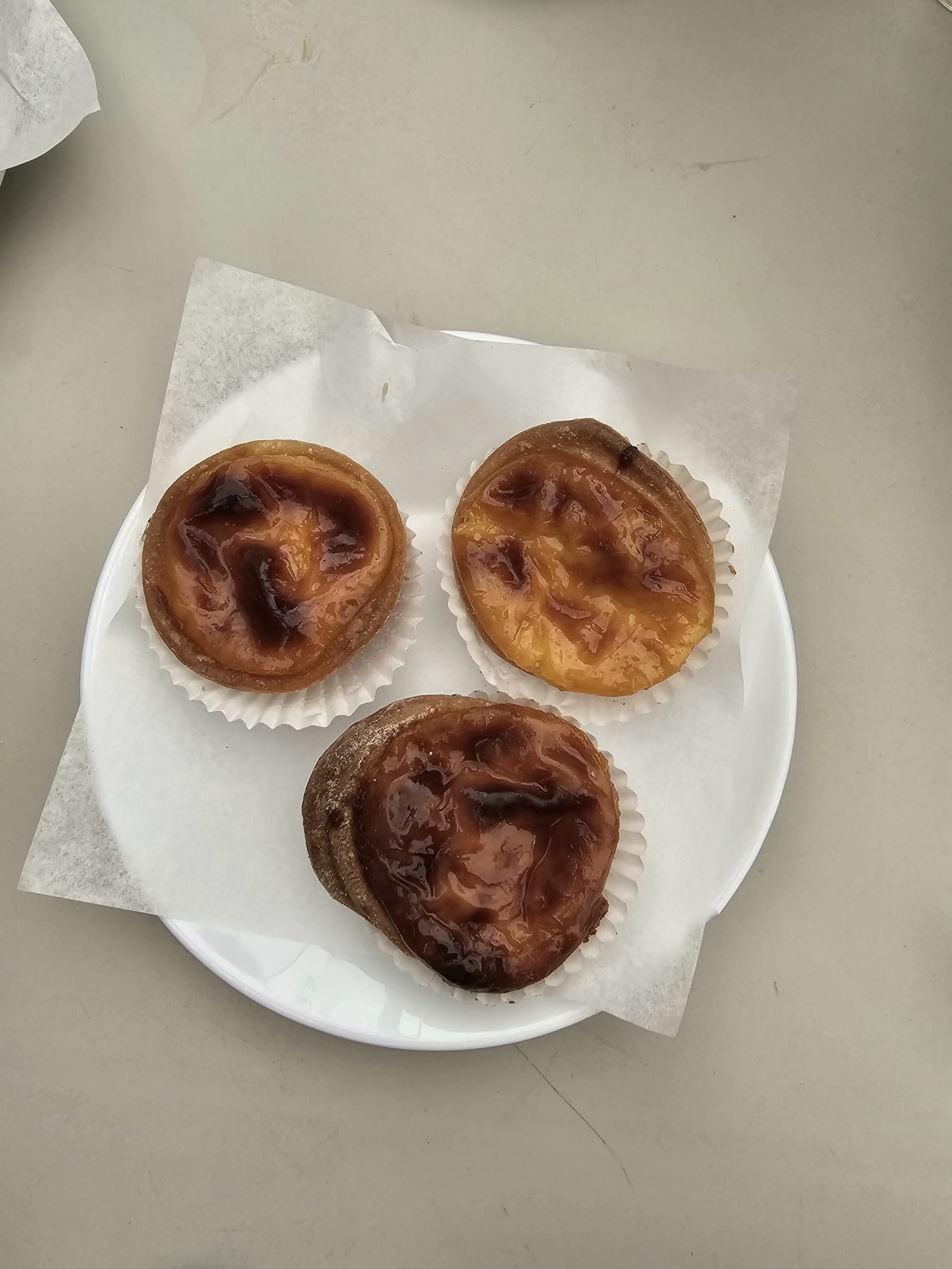 Three Portuguese custard tarts, known as pastéis de nata, are displayed on a white plate with parchment paper, showcasing their golden-brown, caramelized tops.