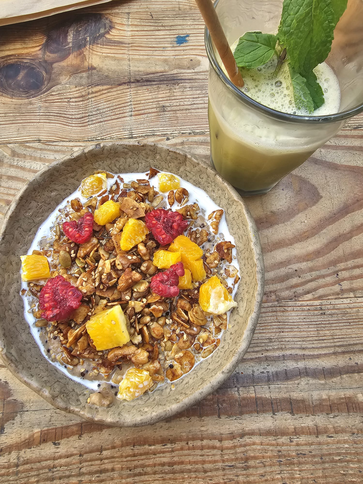 A rustic bowl of granola topped with vibrant raspberries and orange pieces sits on a wooden table, accompanied by a glass of green juice garnished with fresh mint leaves and a straw.